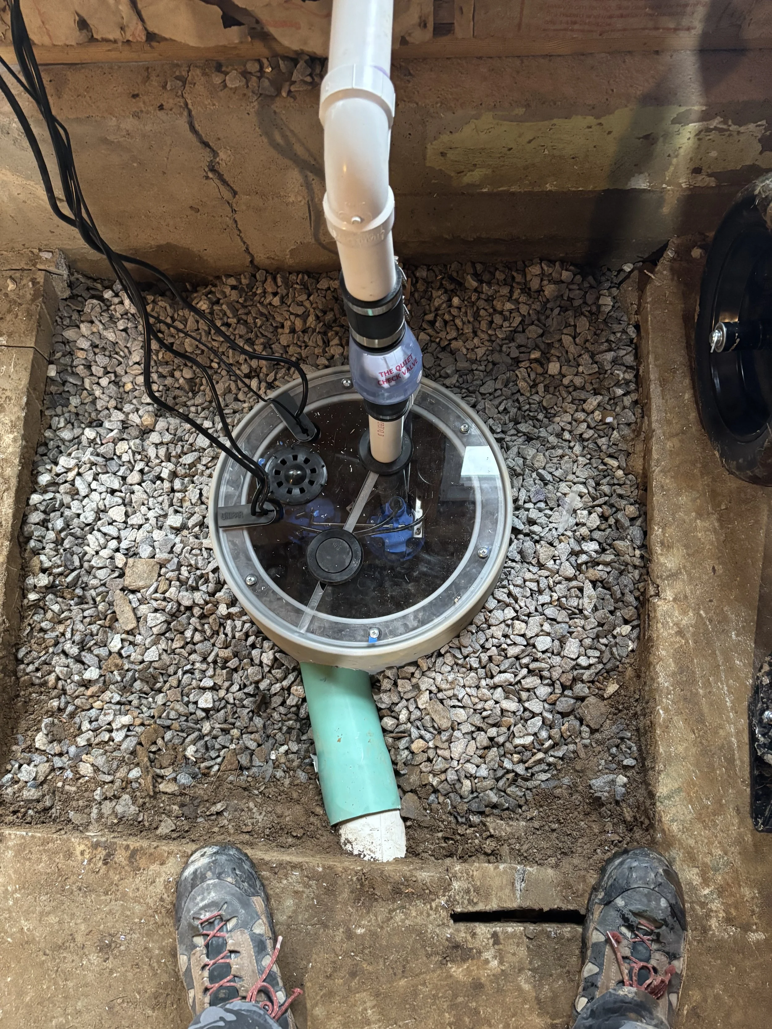 A sump pump installed in a gravel-filled pit with the person’s feet visible in the foreground. basement systems maine bone dry basement turner maine 
