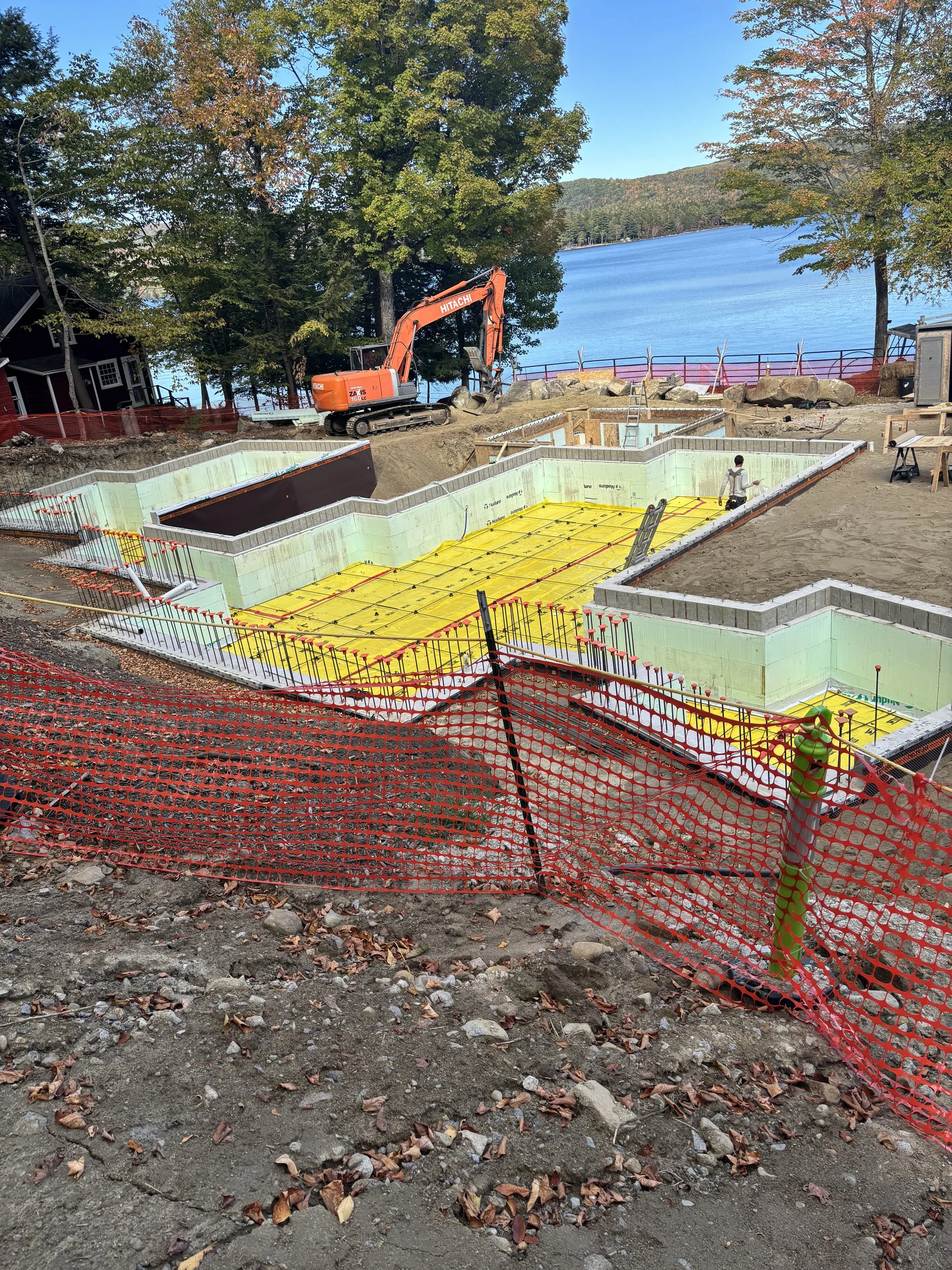 Construction site near a lake with foundation work and an excavator, surrounded by trees with fall foliage. basement systems maine bone dry basement turner maine 