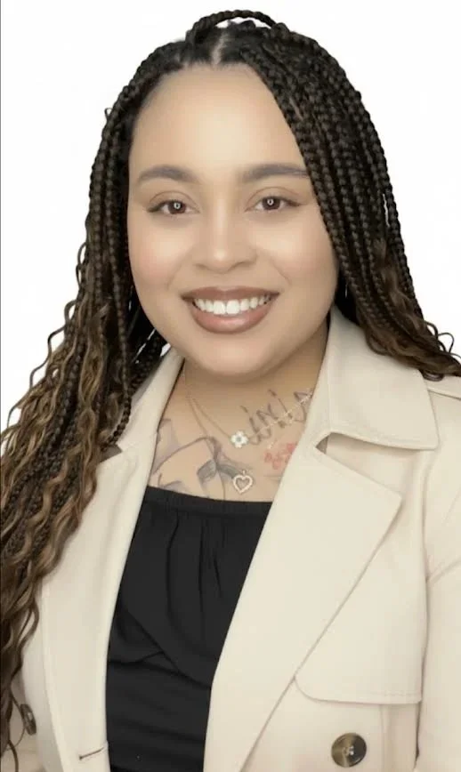 Portrait of a smiling woman with long braided hair, wearing a beige blazer over a black top, with visible tattoos on her chest.