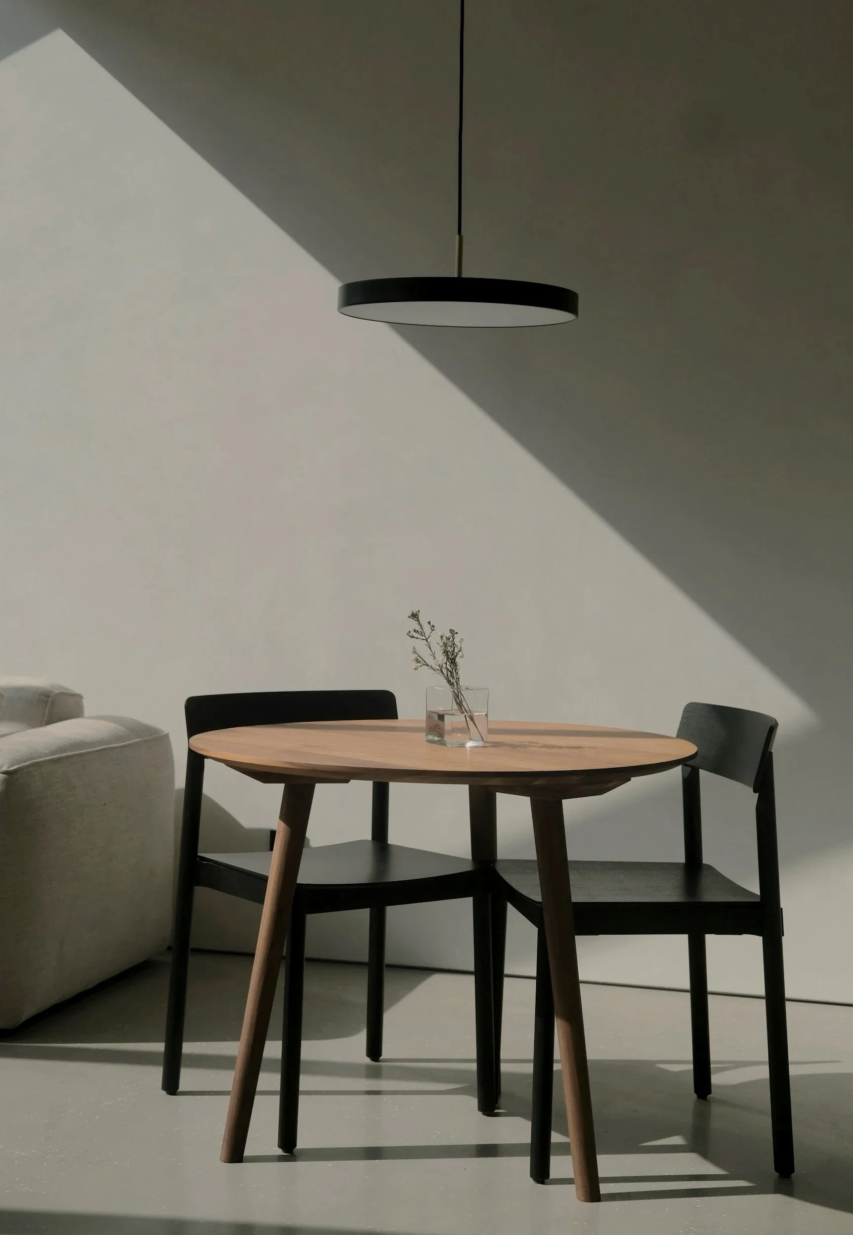 Ein runder Holztisch mit drei schwarzen Stühlen und einer kleinen Vase mit Zweigen. Ein moderner, schwarzer Deckenleuchte hängt über dem Tisch. Der Raum hat weiße Wände und ist minimalistisch gestaltet.