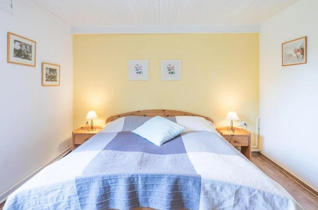 Bett im Schlafzimmer mit zwei Nachttischen und Lesehämtern, gelbe Wand hinter dem Bett, weiße Wände seitlich, Bilder an den Wänden, kleine Nachttischlampen, Tagesdecke in Blau, Grau und Weiß, einzelne Kissen auf dem Bett.