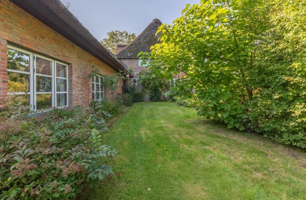 Garten mit grünem Rasen, Büschen und Bäumen neben einem Backsteinhaus mit weißen Fenstern.