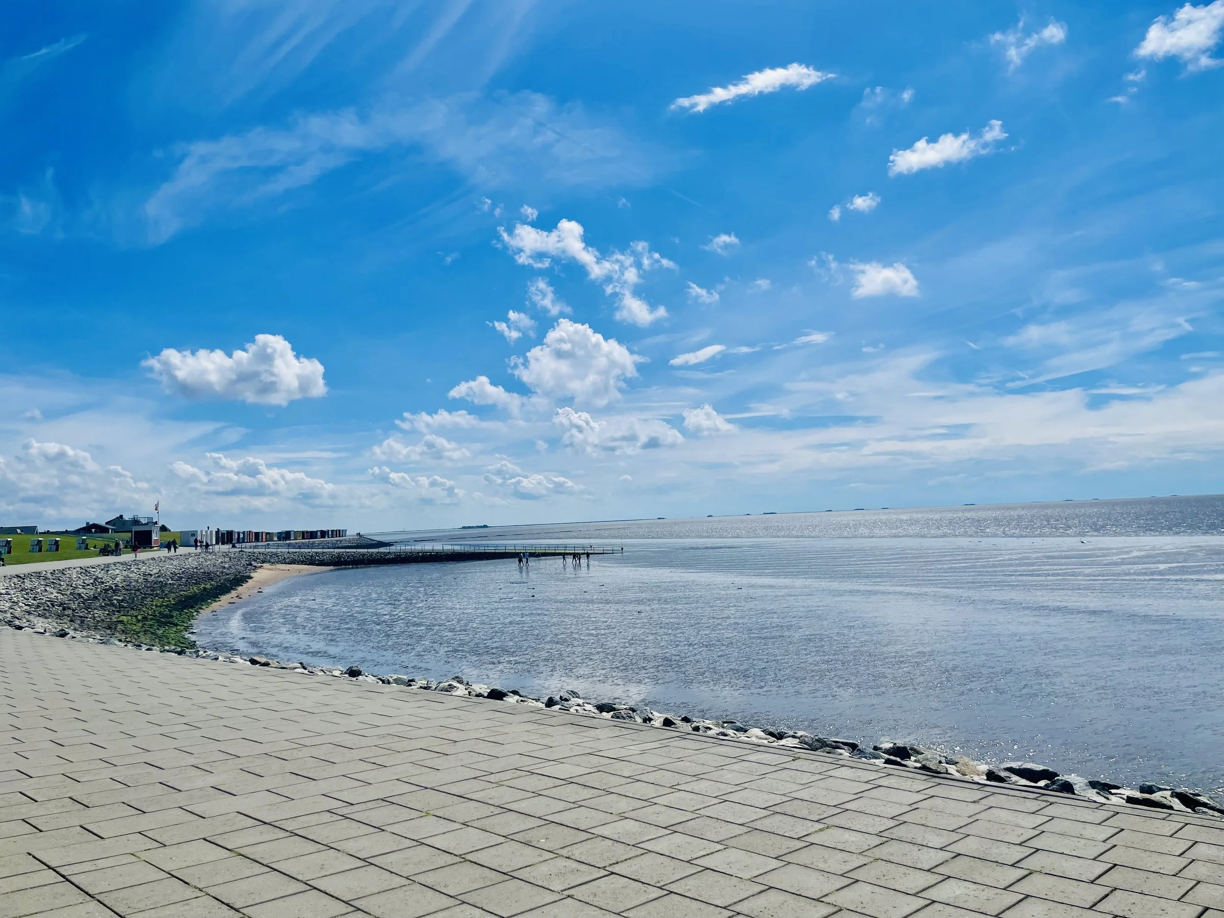 Ein Strand mit einem gepflasterten Weg, der an das Wasser grenzt, bei sonnigem Himmel mit Wolken, im Hintergrund sind einige Gebäude und ein Hafen zu sehen.