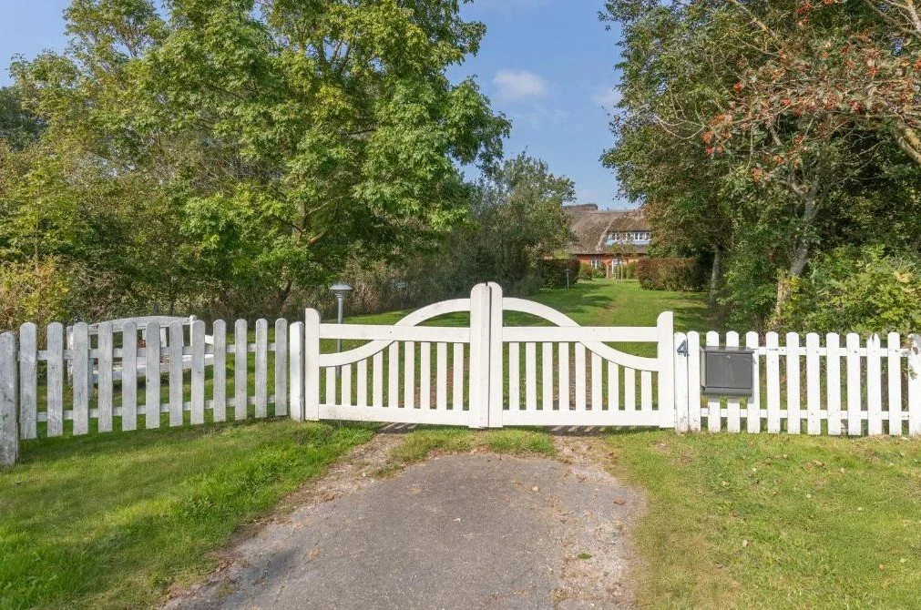 Ein weißer Gartenzaun mit Tor, umgeben von grünen Bäumen und einem gepflegten Garten, im Hintergrund ein Haus mit Dach und Fenstern, bei sonnigem Himmel.