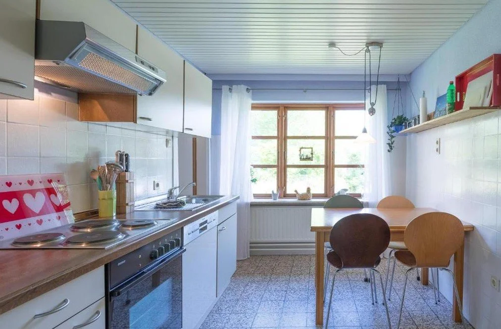 Küche mit weißen Schränken, Holzarbeitsplatte, Herd, Spüle, Fenster, Esstisch mit vier Stühlen, Dekorationsgegenständen und Geschirr.