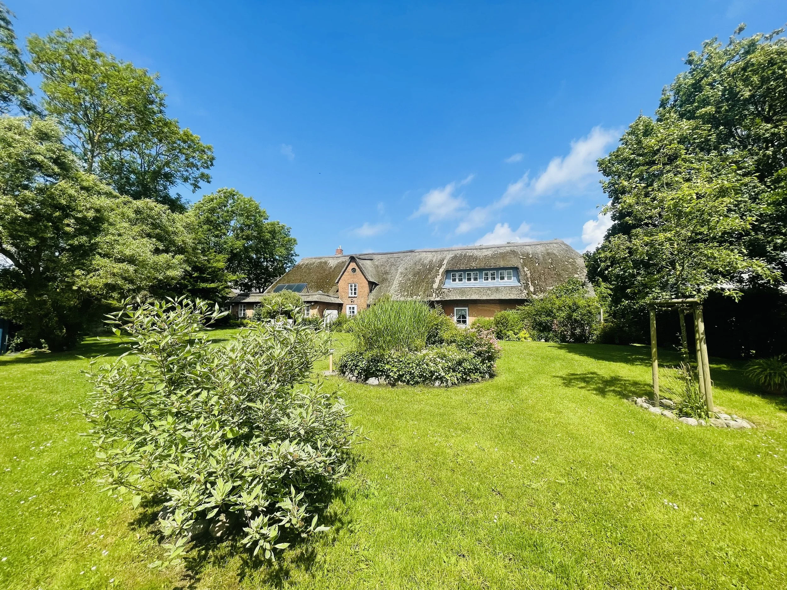 Ein großes, altes Haus mit Reetdach in einem schönen, grünen Garten, umgeben von Bäumen, bei sonnigem Himmel.