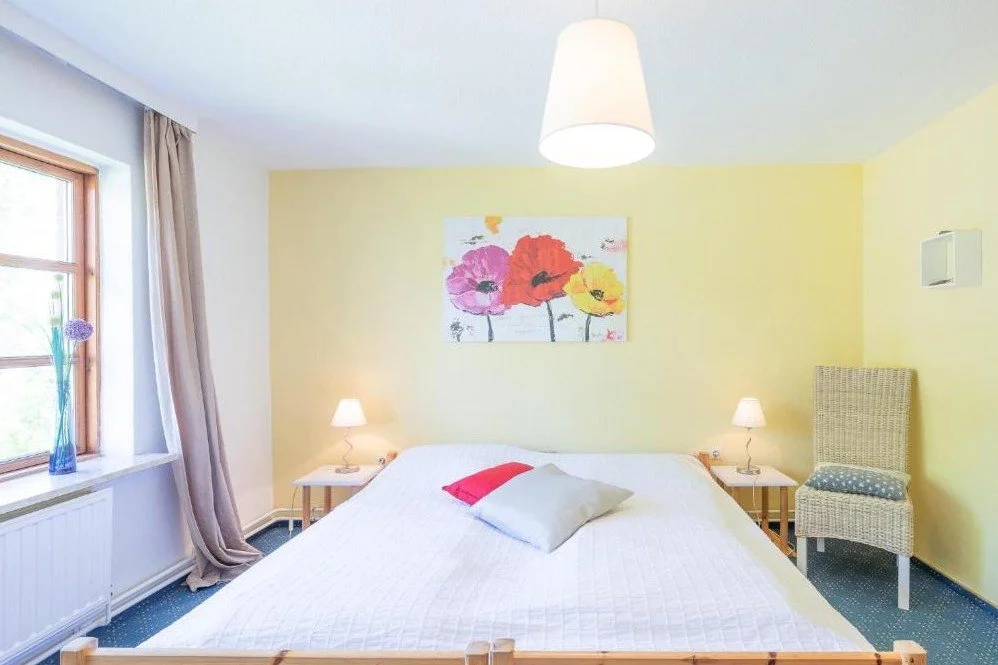 Schlafzimmer mit Doppelbett, Wandbild mit bunten Blumen, Nachttische mit Lampen, Fenster mit Vorhängen, Wanduhr, Rattanstuhl, hellgelbe und weiße Wände.