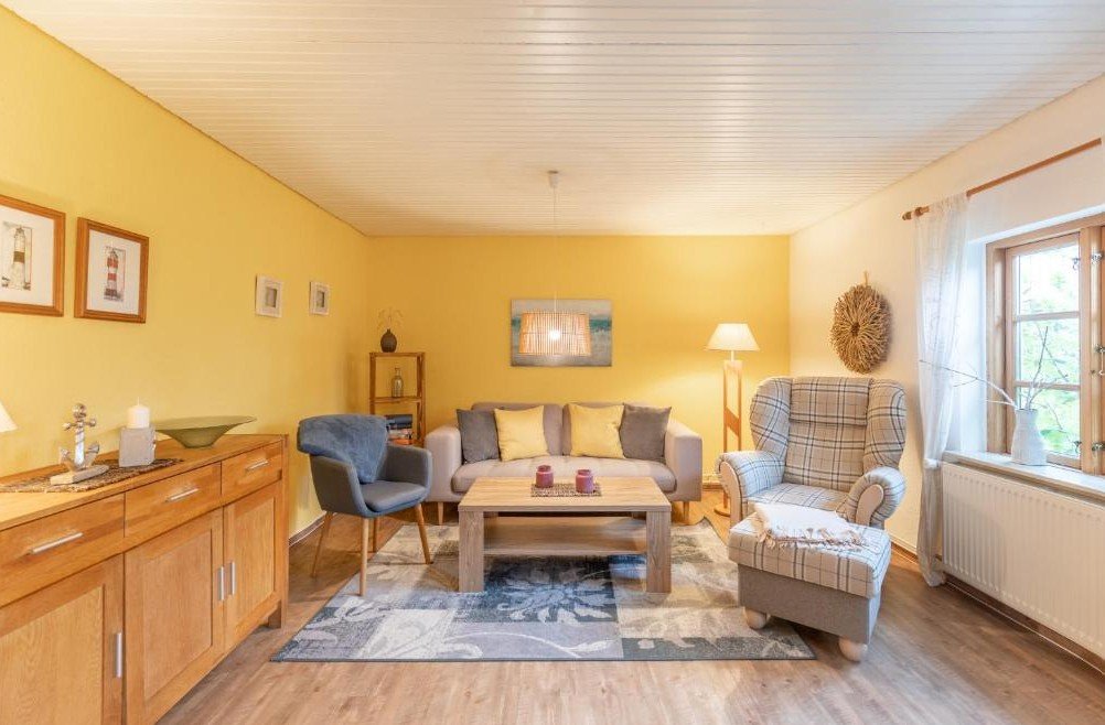 Gemütliches Wohnzimmer mit gelber Wand, hellem Holzfußboden, grauer Sofa mit gelben und grauen Kissen. Großer Sessel mit Karo-Muster, Stehlampe, Fenster mit weißen Vorhängen, Dekorationen an der Wand, Couchtisch und einem Sideboard.