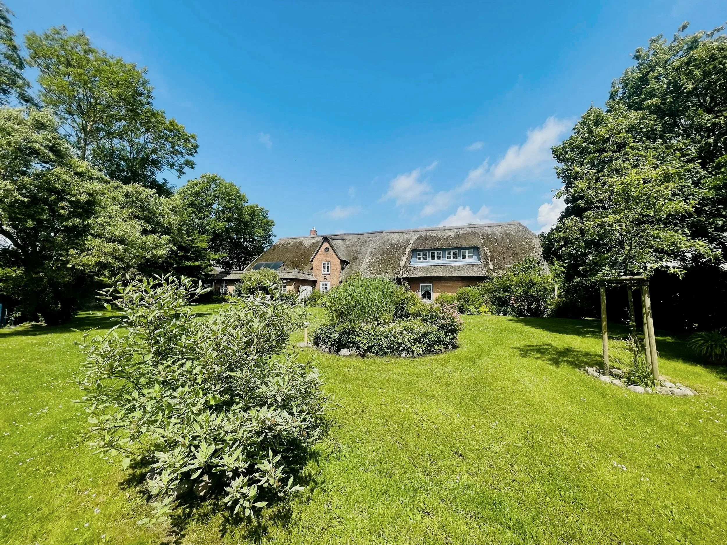 Ein landliches Haus mit reetgedecktem Dach, umgeben von grünen Bäumen, gepflegtem Garten und blühenden Sträuchern unter blauem Himmel.