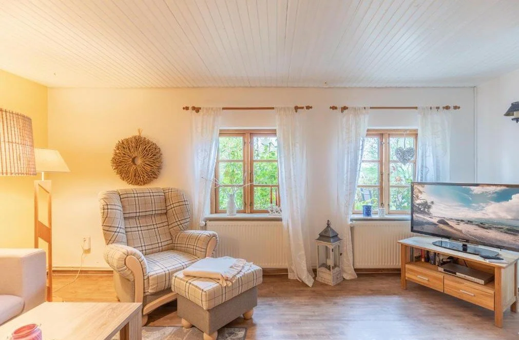 Gemütliches Wohnzimmer mit Sessel, Fernseher und großen Fenstern mit weißen Vorhängen.