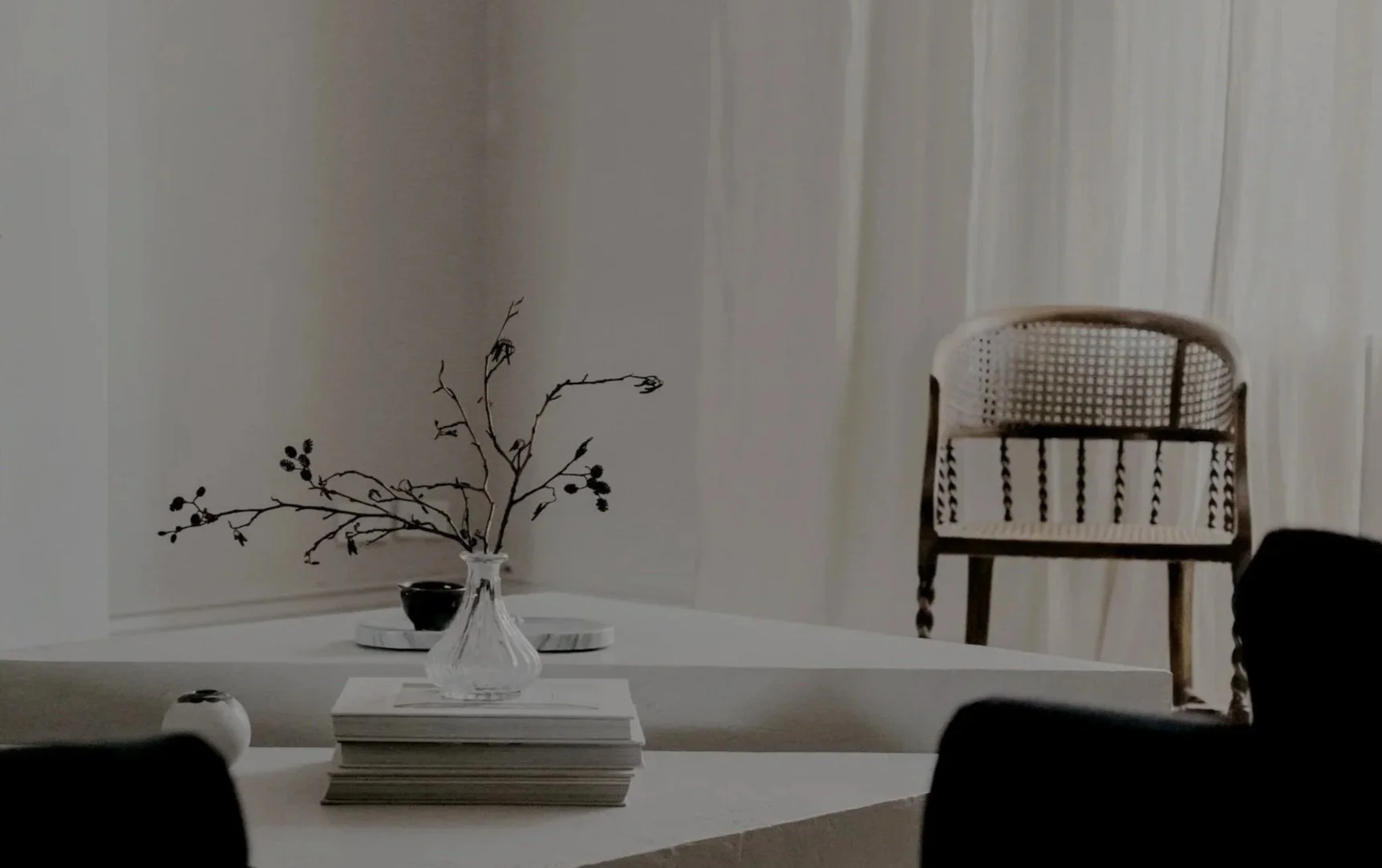 Innenaufnahme eines minimalistisch eingerichteten Raumes mit weißem Tisch, auf dem eine Vase mit Zweigen steht, und einem Holzstuhls im Hintergrund.