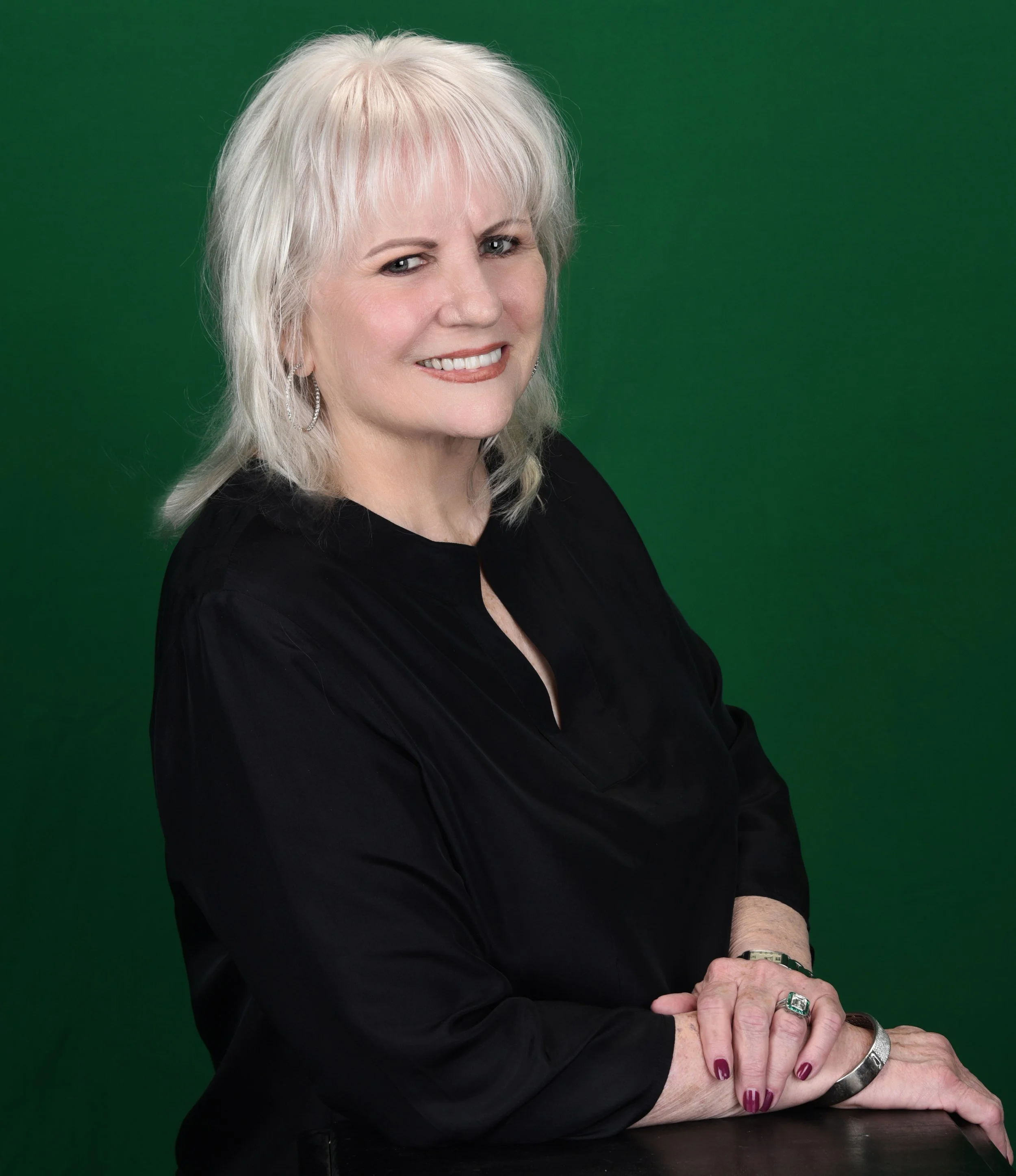A smiling woman with shoulder-length white hair posing in front of a green background, wearing a black top and jewelry.