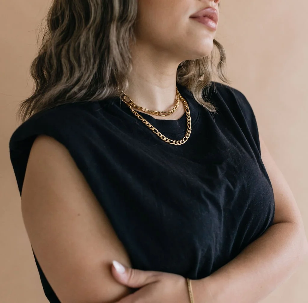Close-up of a woman wearing layered gold chain necklaces and a black sleeveless top, with her arms crossed.