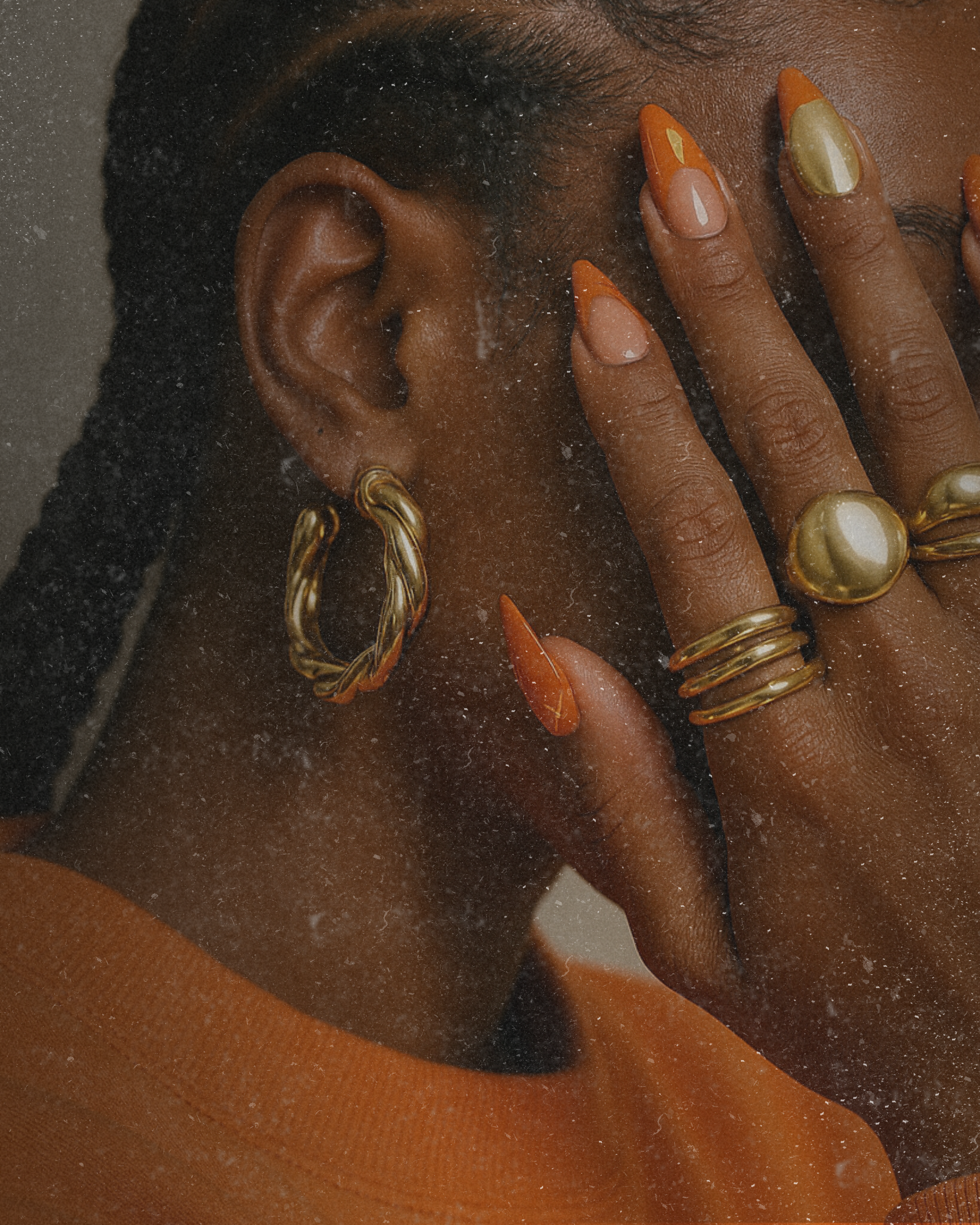 Close-up of a woman with gold jewelry and orange and metallic nail polish, touching her face and ear.