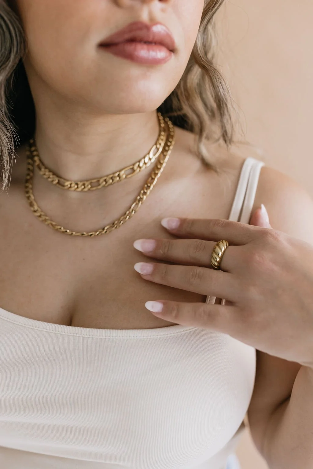 Close-up of a woman wearing layered gold necklaces, a gold ring, and a white tank top, with her hand resting on her shoulder.