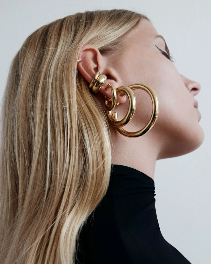 A woman with long blonde hair wearing multiple large gold hoop earrings and a black turtleneck.