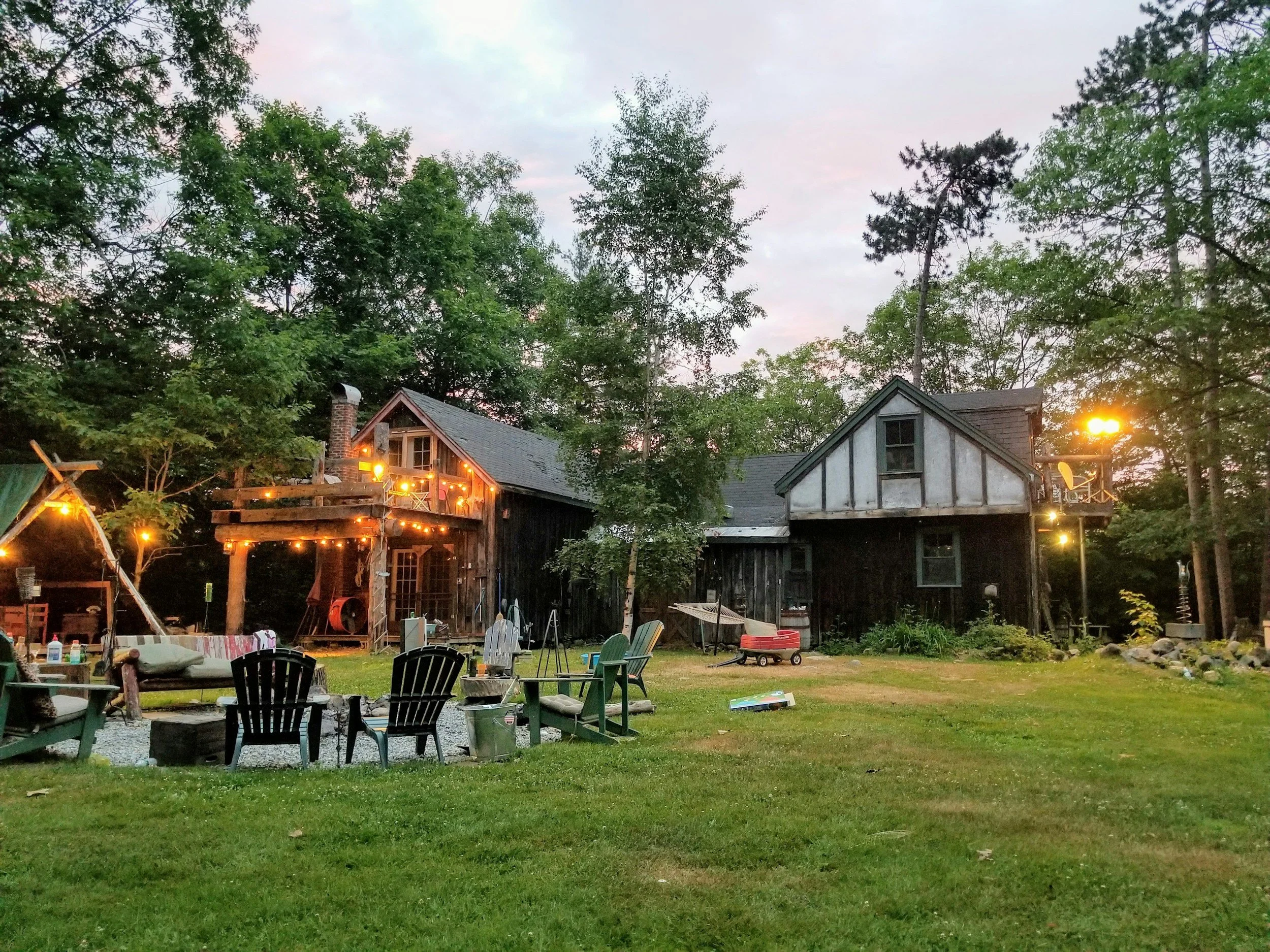 A rustic backyard scene at dusk with a two-story black wooden house, string lights, outdoor chairs, and trees surrounding the area.