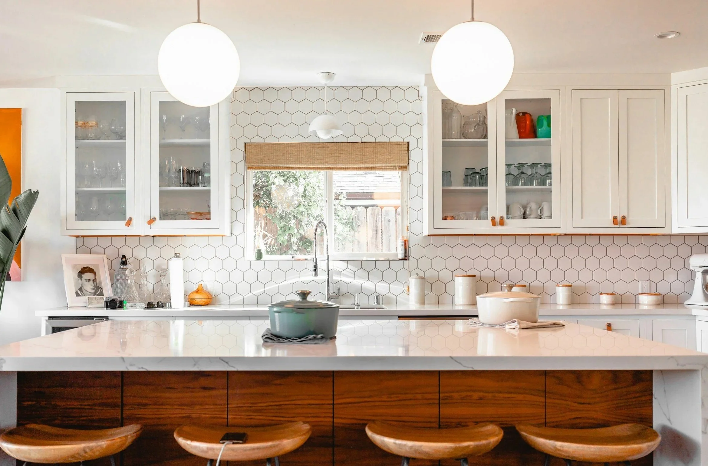 Bright kitchen with white cabinets, a central island with a marble countertop, and a window above the sink. Decor includes glassware, jars, and framed photo, with pendant lights hanging from the ceiling.