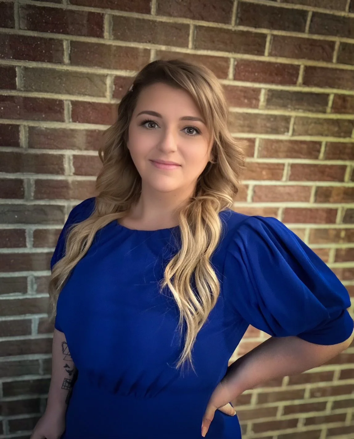 A woman with long, wavy blonde hair, wearing a royal blue dress with puffed sleeves, standing against a brick wall. She has a slight smile and is looking at the camera.