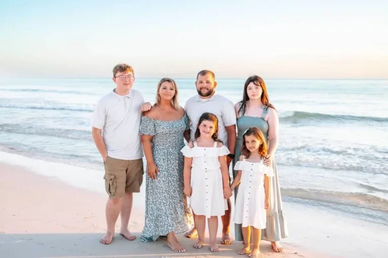 Family of six standing on the beach near the ocean, smiling, with sunset sky in the background.