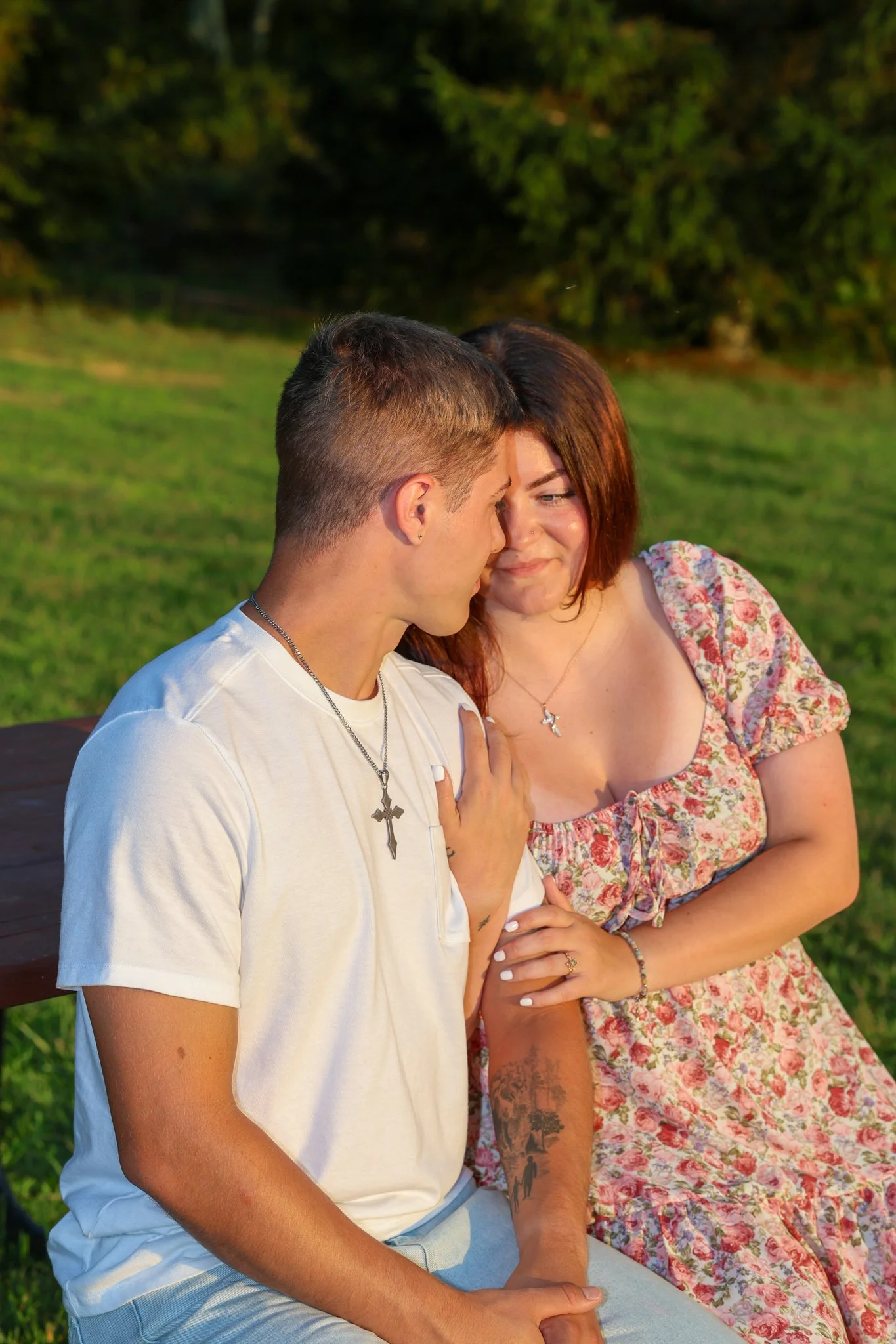 A young man and woman sitting close together outdoors, touching foreheads and gazing at each other during sunset in a grassy field with trees in the background.