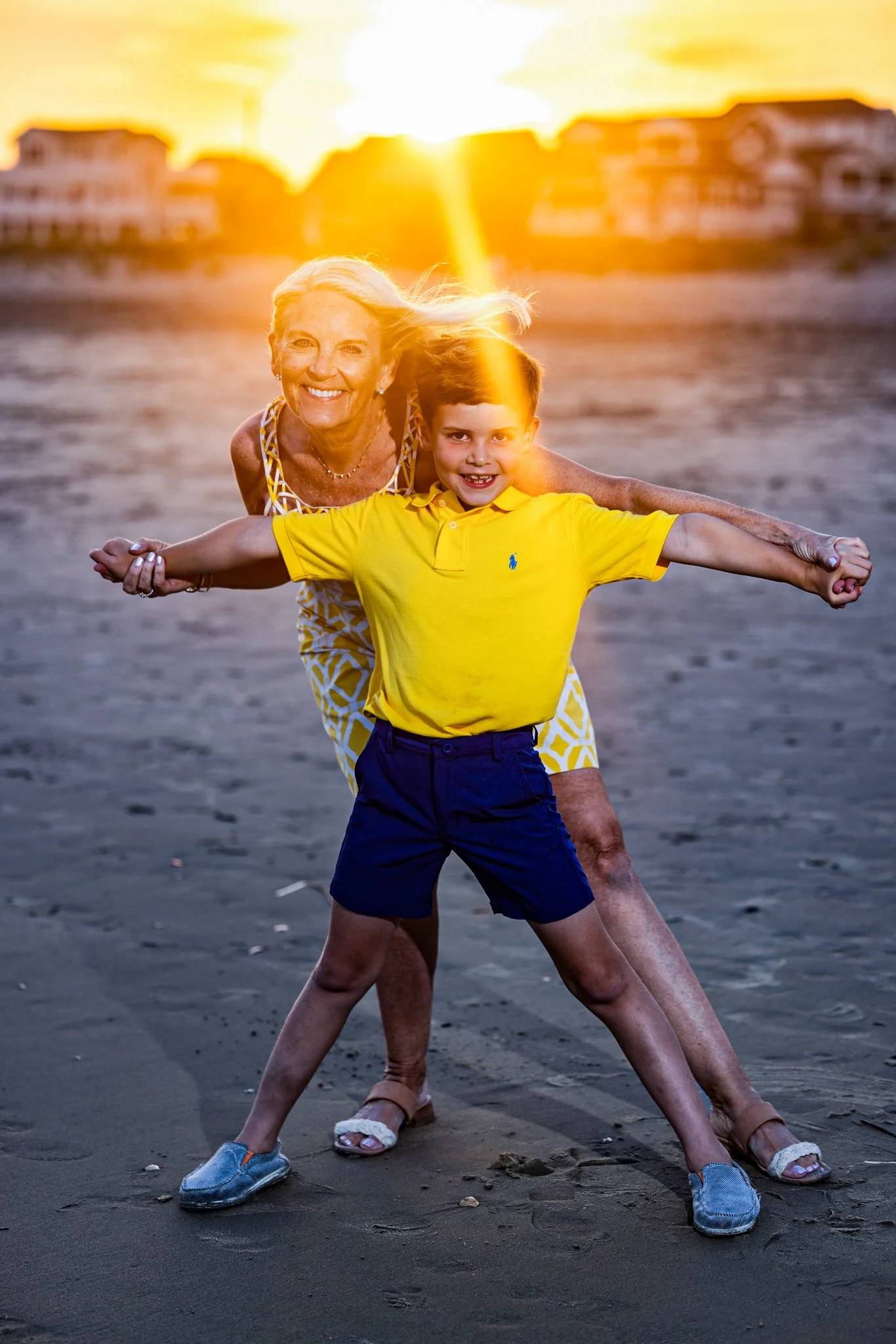 An elderly woman and a young boy smiling and holding hands on a beach at sunset, with buildings in the background.
