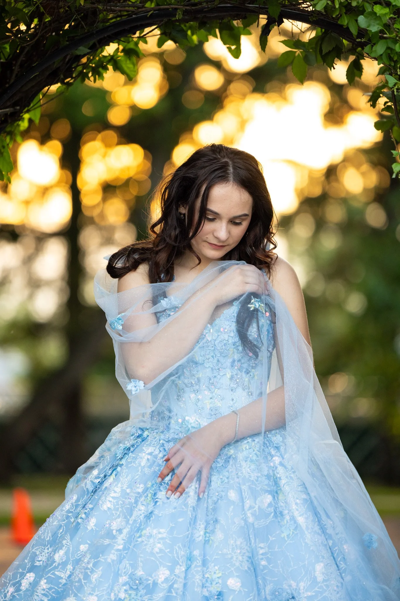 A young woman in a light blue, intricately embroidered dress with a sheer shawl, standing outdoors under a tree with green leaves at sunset, looking down with a gentle expression.