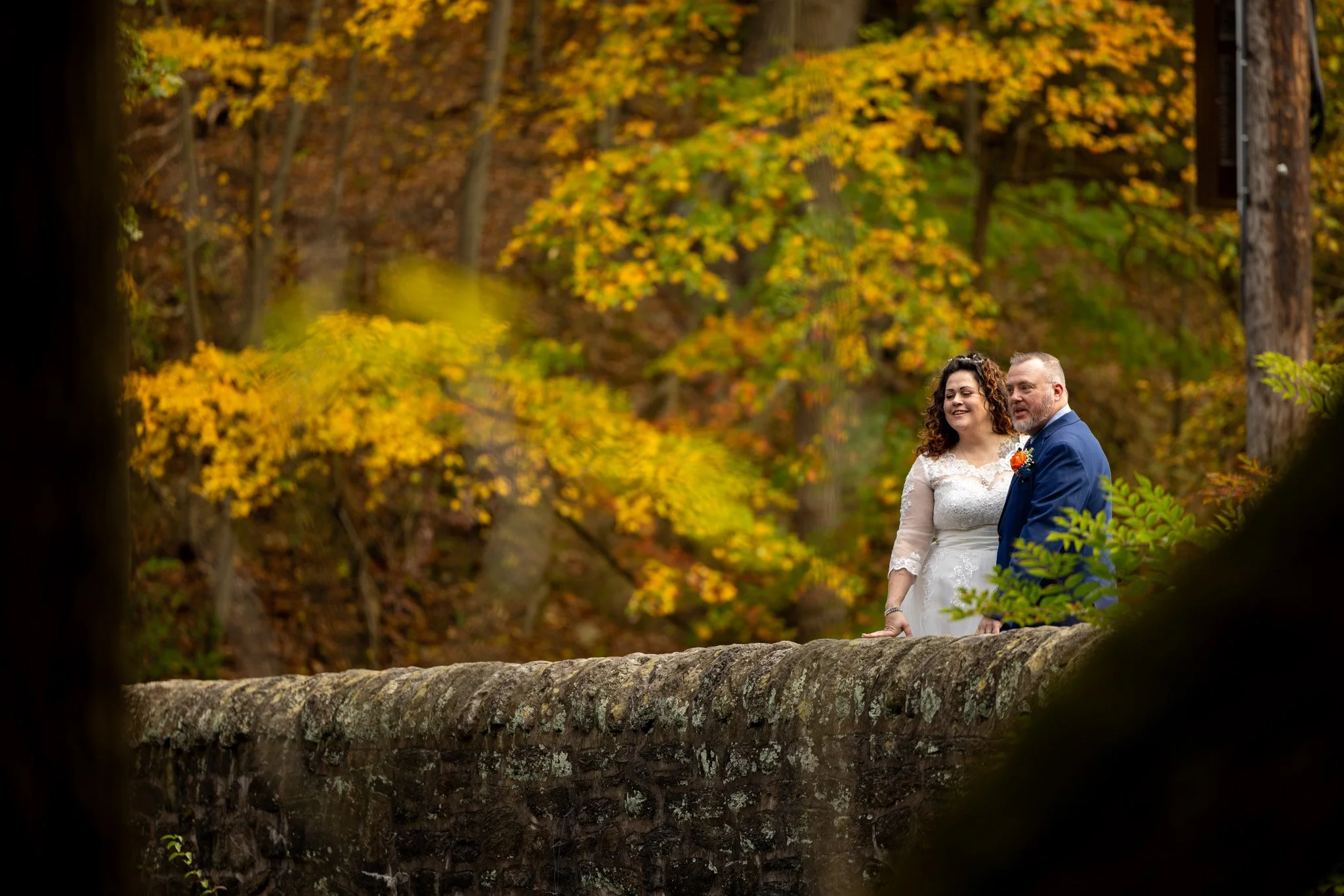 A newlywed couple stands on a stone bridge in a forest with autumn foliage, smiling and looking towards the distance.