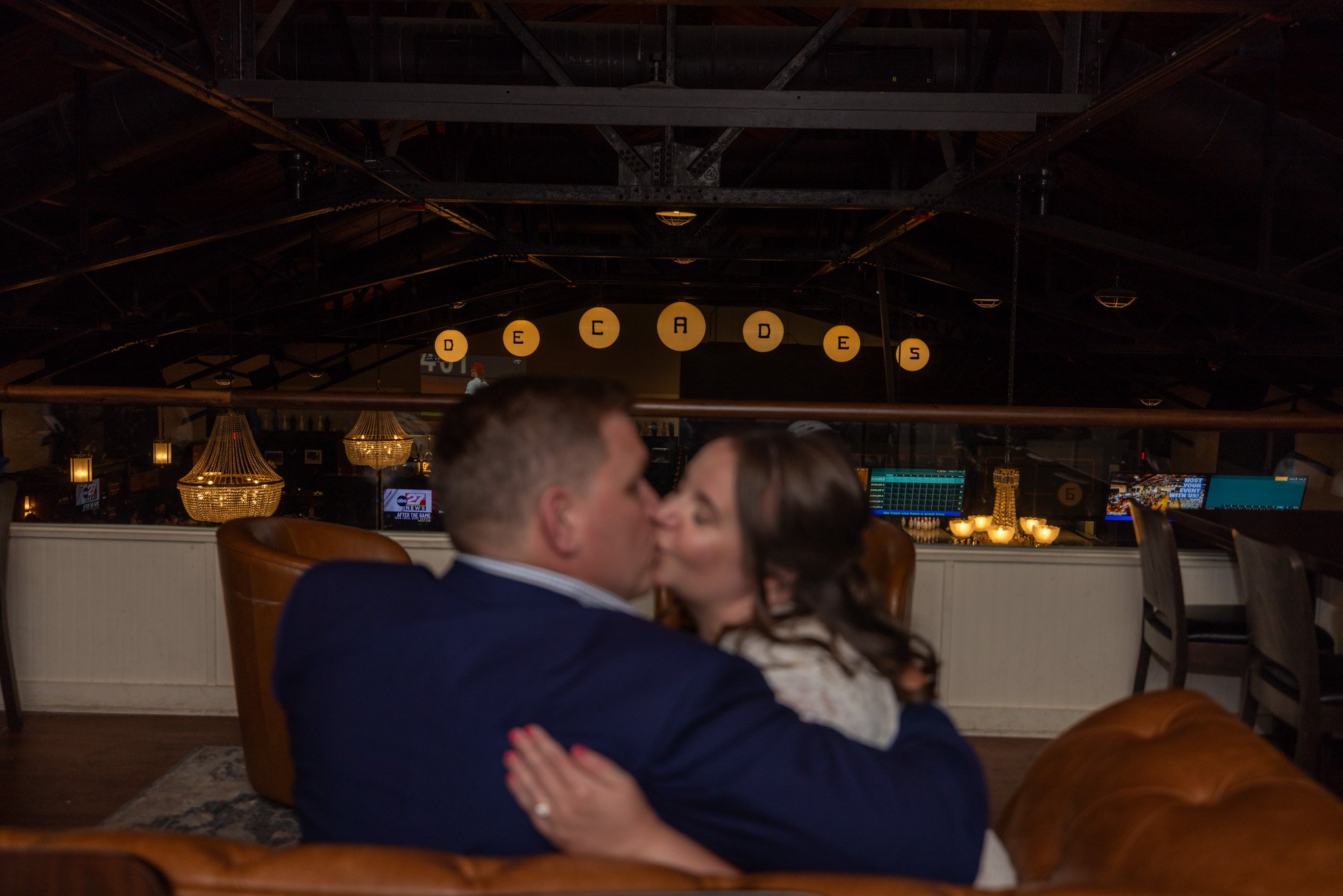 A couple sharing a kiss while sitting on a brown leather sofa in a dimly lit room with hanging lights and a dark ceiling.