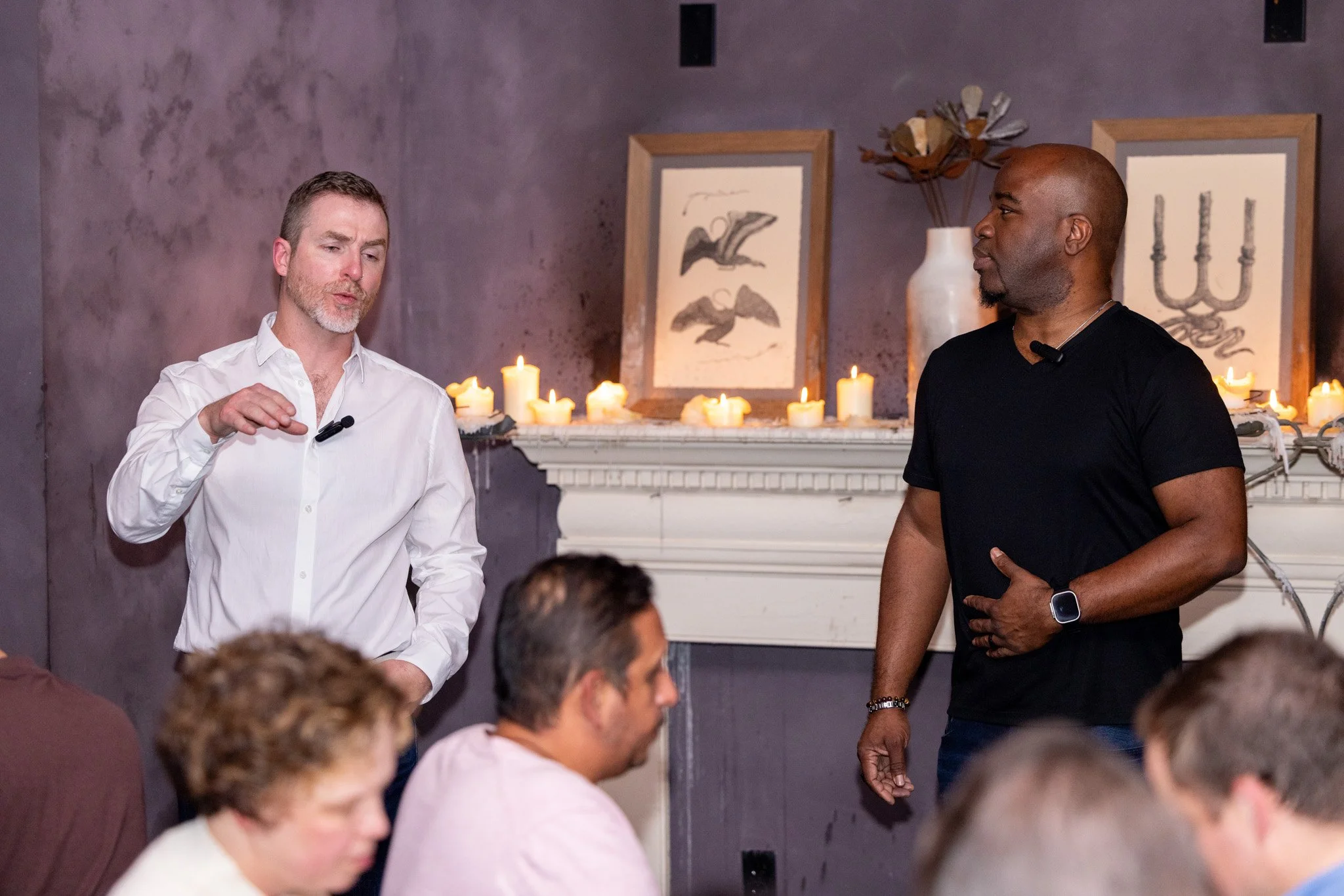 Two men are standing and speaking in front of a seated audience. One man is wearing a white shirt and the other is wearing a black t-shirt. Behind them is a fireplace with lit candles and framed artwork on the wall.