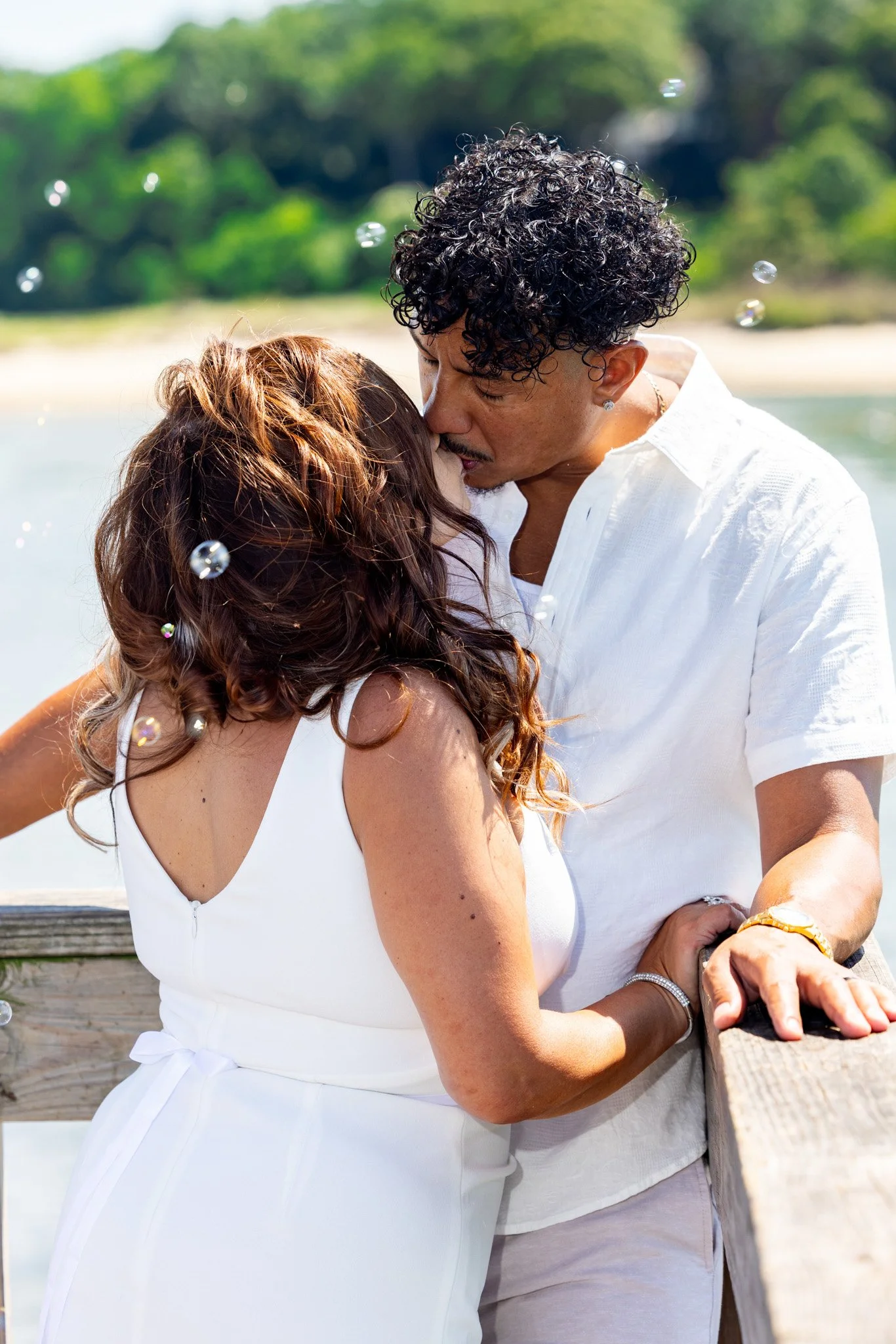A couple kissing outdoors near a body of water, with a background of green trees and a sandy shoreline, surrounded by floating bubbles.