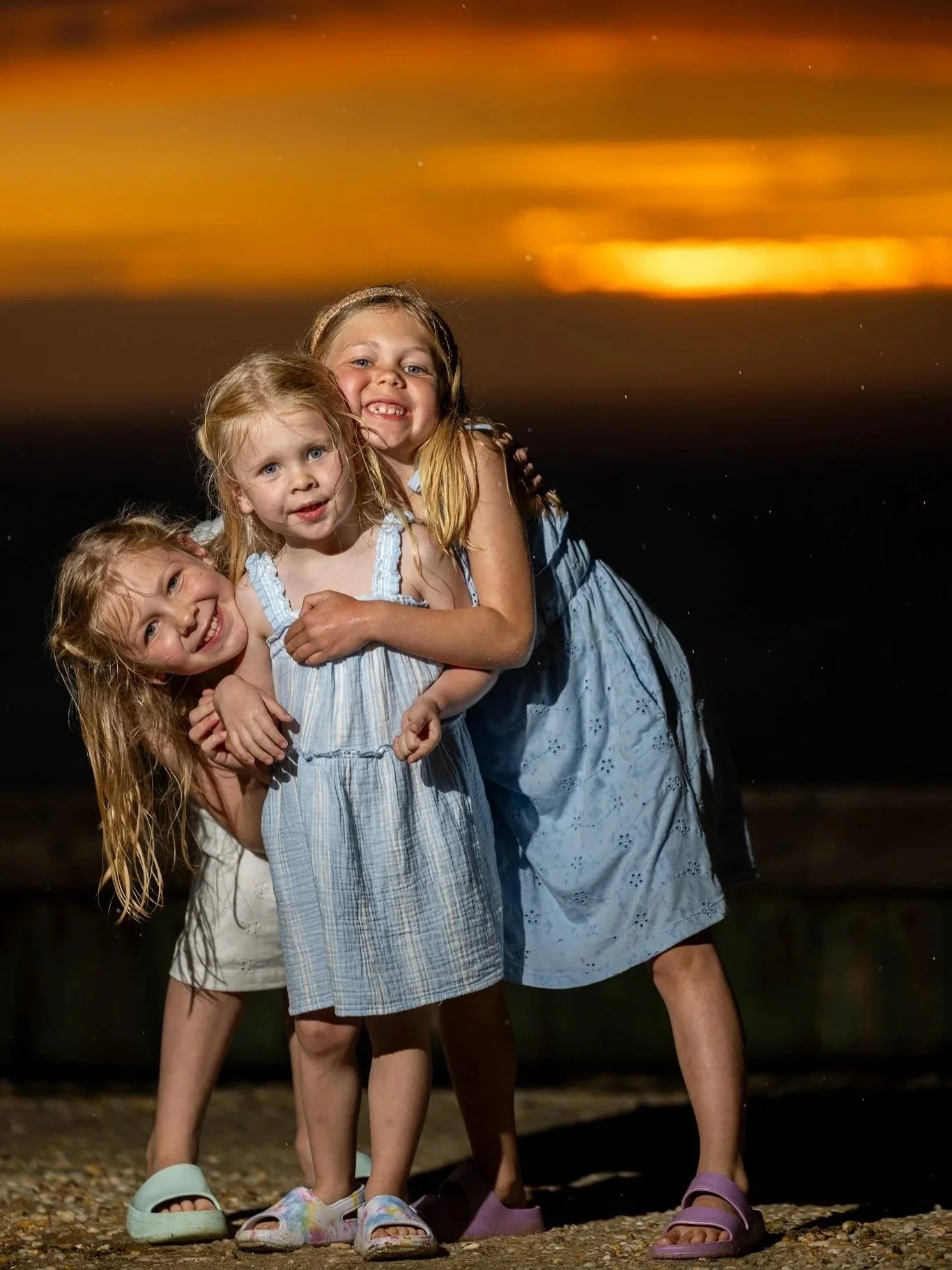 Three young girls with wet hair and wearing summer dresses, smiling and hugging each other on a beach at sunset.