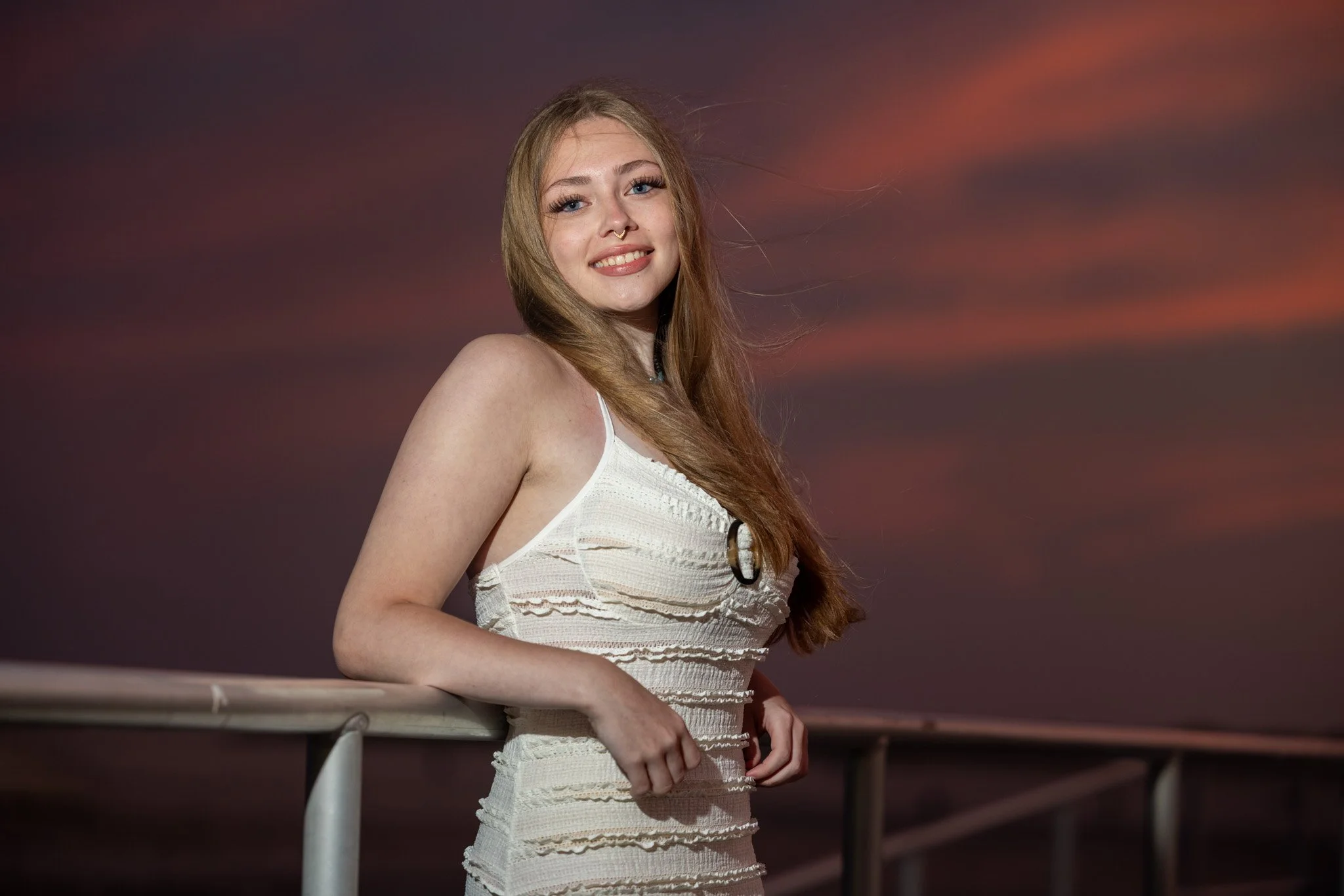 A young woman with long reddish-brown hair, wearing a white dress, smiling and leaning on a railing outdoors during sunset with pink and purple clouds in the background.