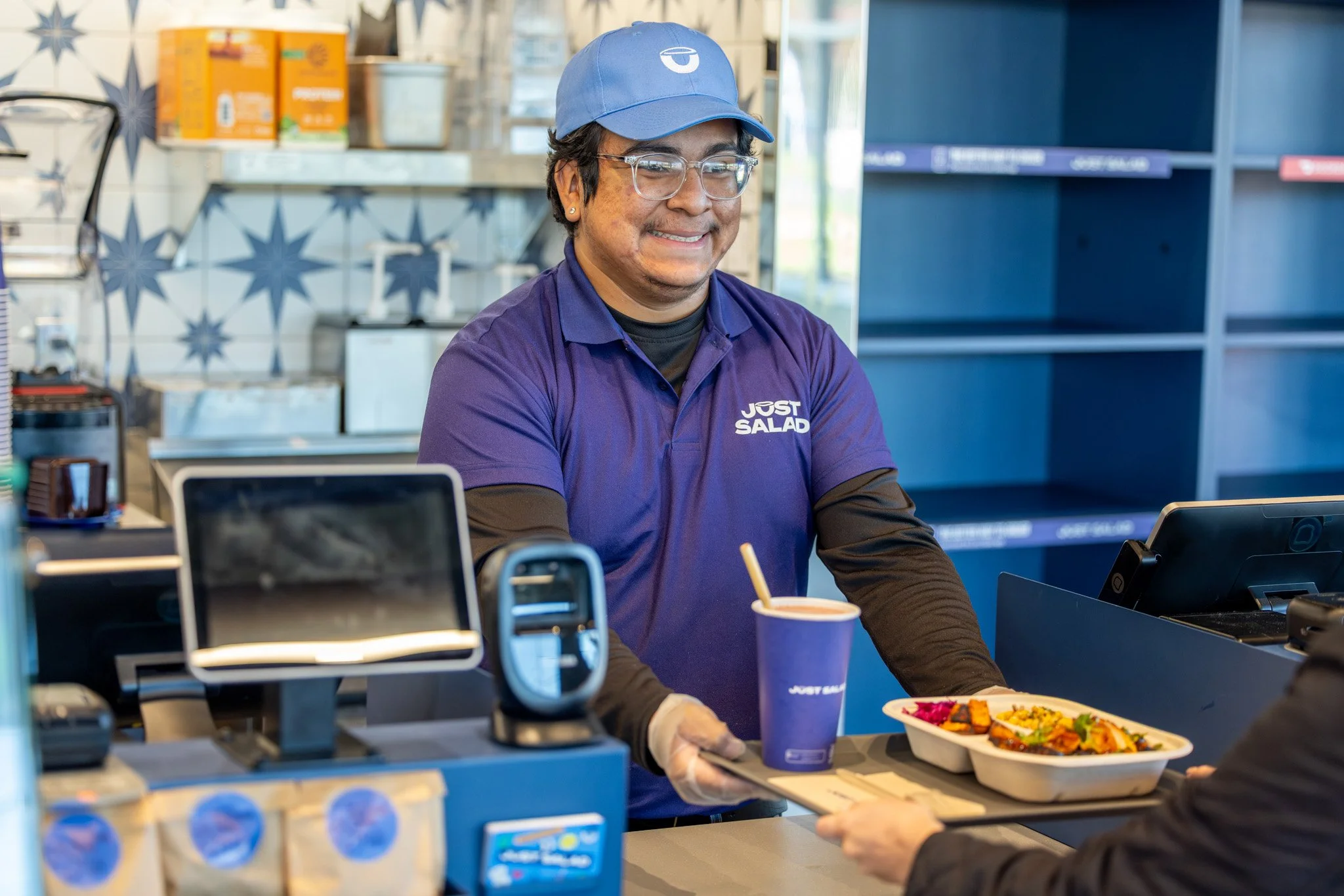 A smiling fast-food worker in a purple uniform and blue cap serving a meal at a counter in a restaurant. The worker is holding a tray with a food container and a drink cup, and is handing it to a customer.