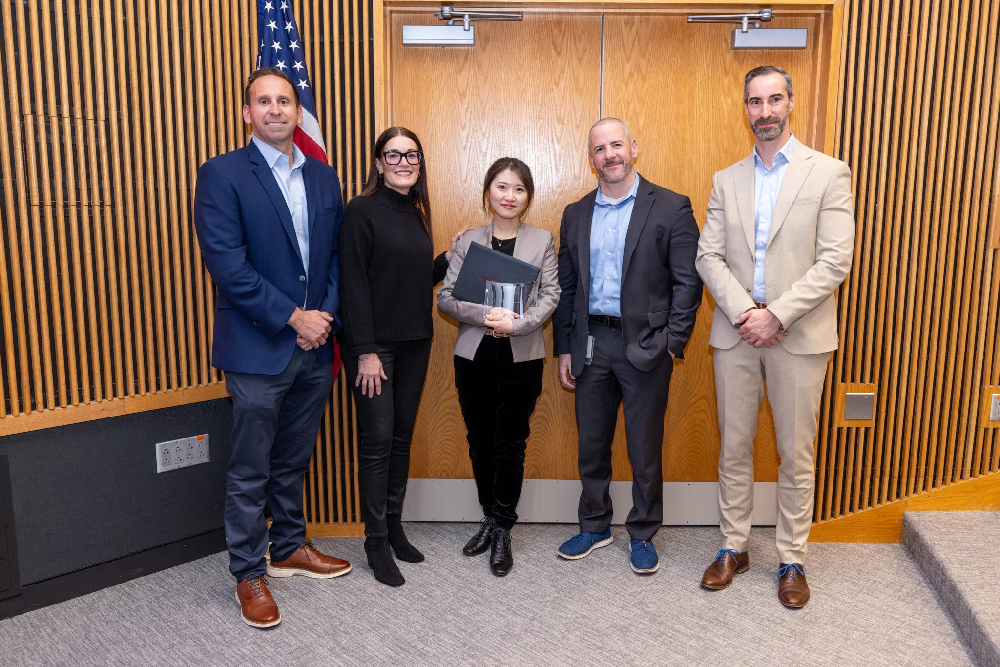 Group of five people standing in a line in an indoor setting, with a woman holding a diploma and a graduation cap, suggesting a graduation ceremony.