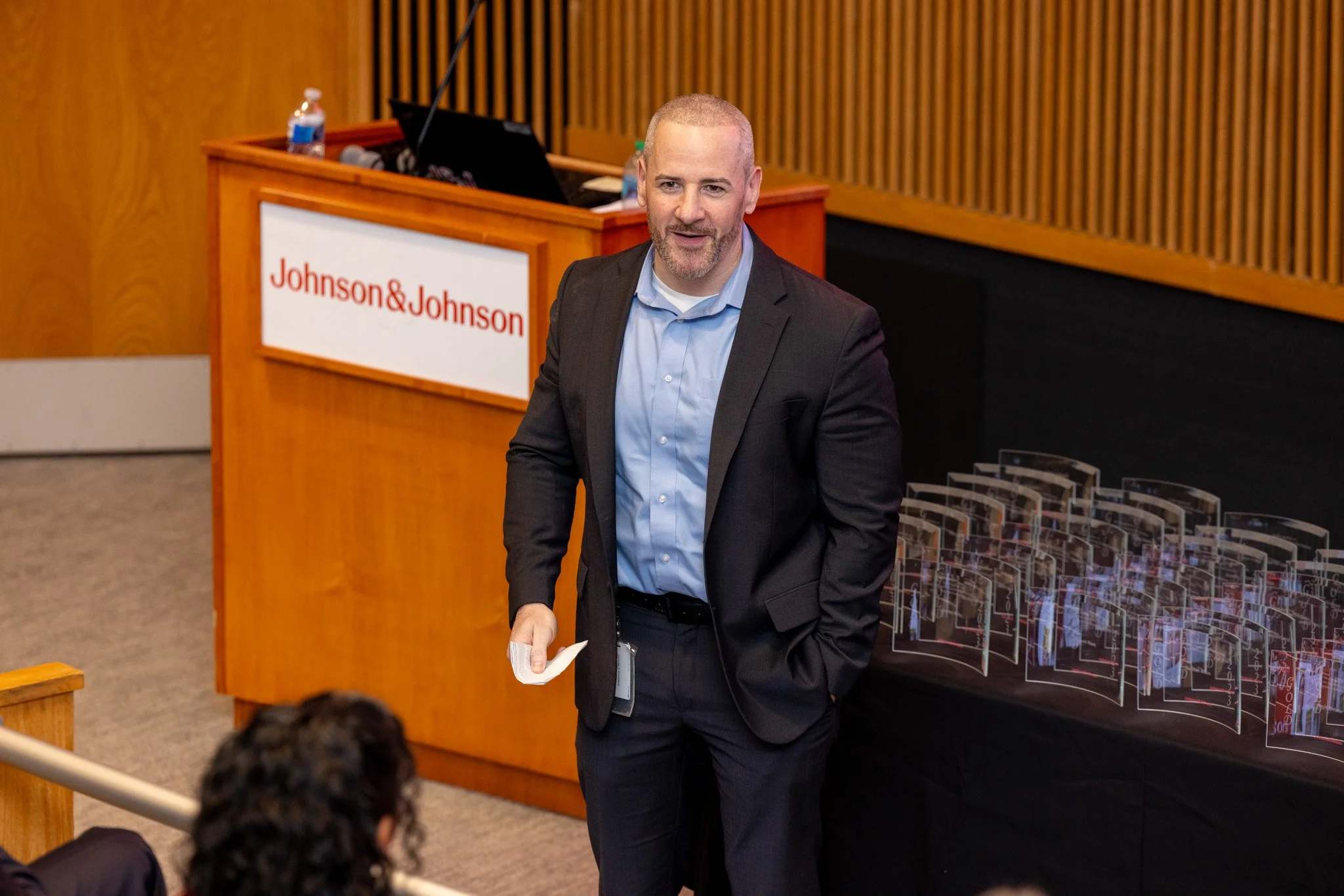 A man in a suit holding a piece of paper, standing in front of a Johnson & Johnson podium, speaking to an audience in a conference room.