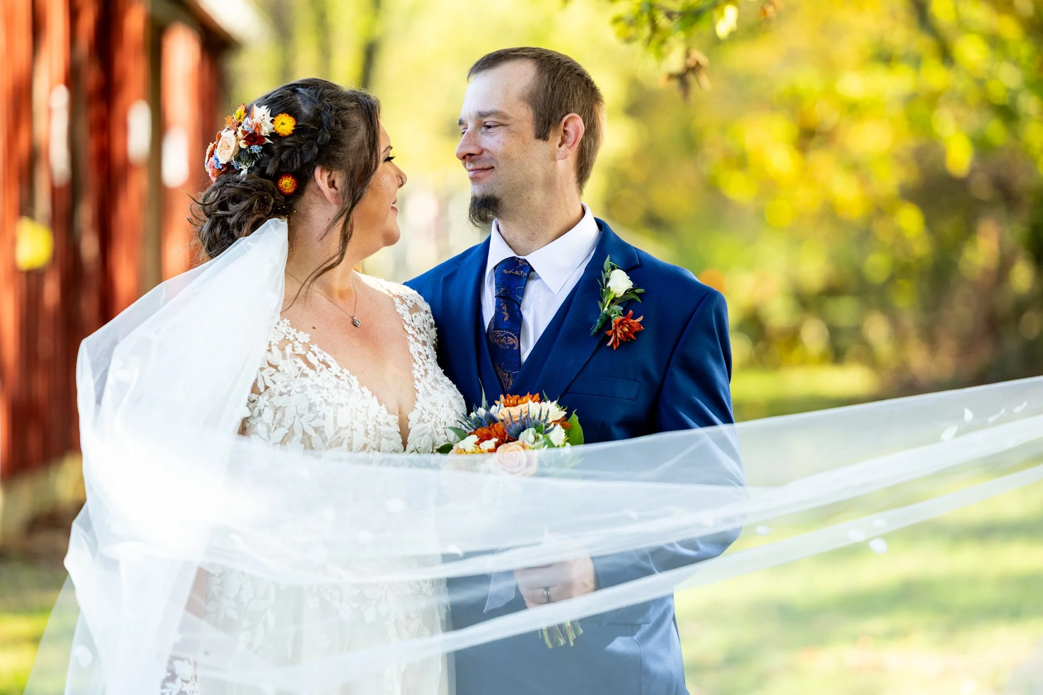 A bride and groom looking at each other outdoors with trees and greenery in the background. The bride wears a lace wedding gown and a floral hairpiece, while the groom wears a blue suit with a tie and a boutonniere. The bride holds a colorful bouquet