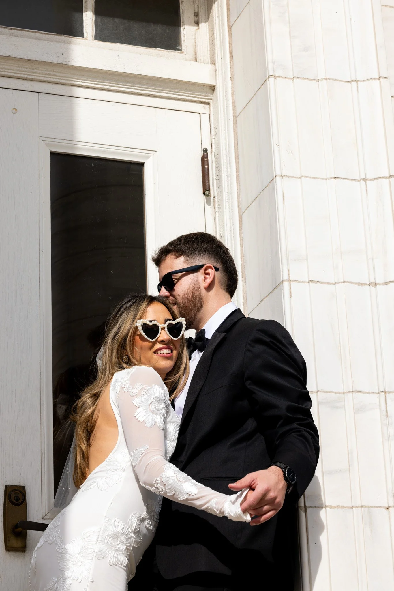 Groom kissing the bride's head as she wears heart-shaped sunglasses in a fashion-inspired wedding portrait by David Pierre Studio