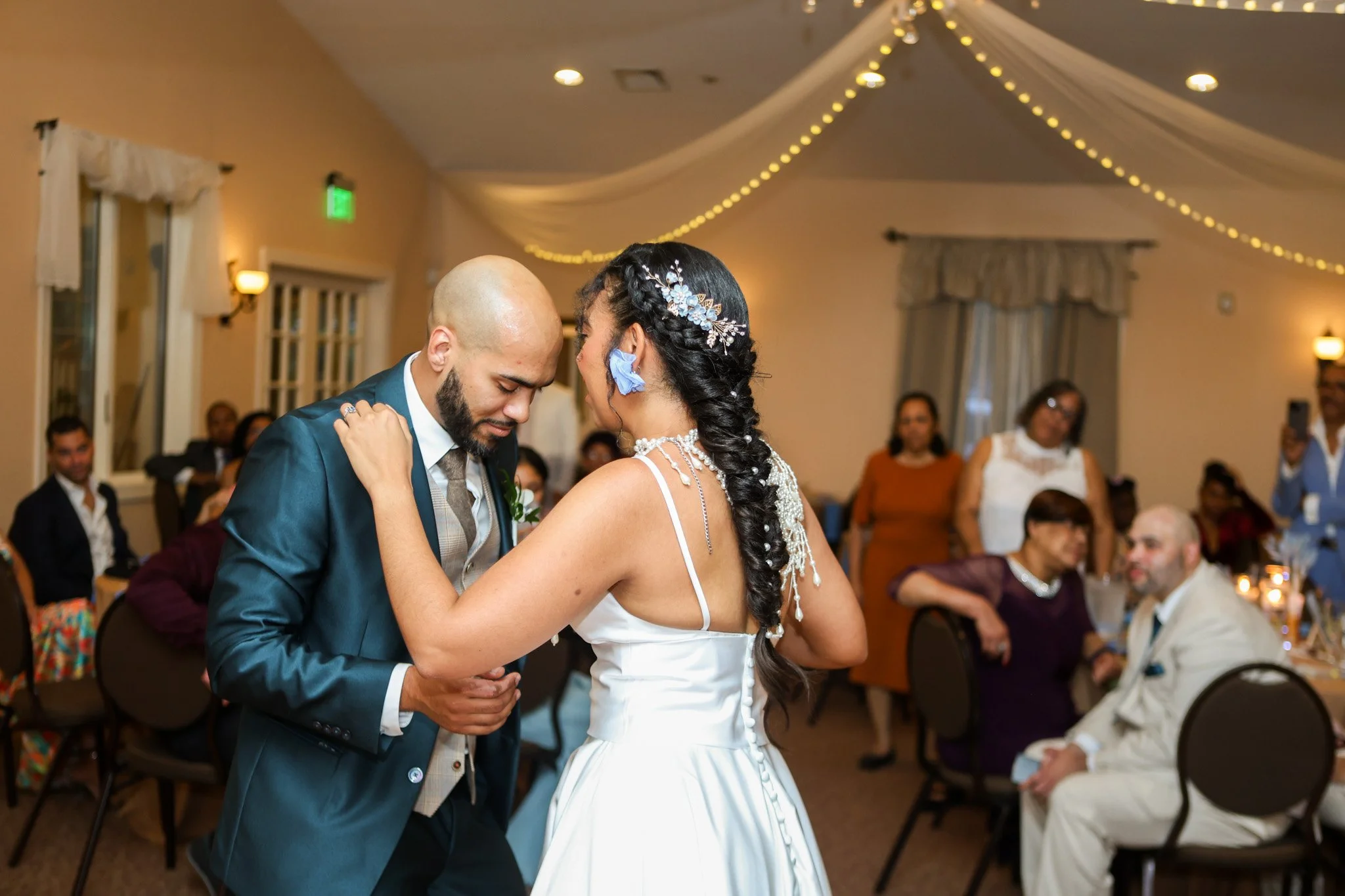 Bride and groom sharing their first dance as guests watch in a reception hall with string lights, captured by David Pierre Studio
