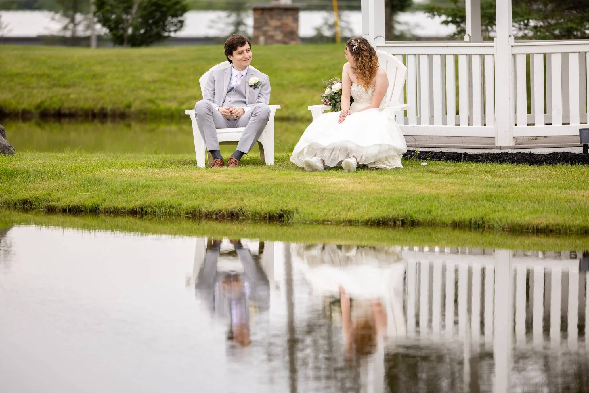 Bride and groom sitting in white chairs beside a pond with their reflection in the water, captured by David Pierre Studio