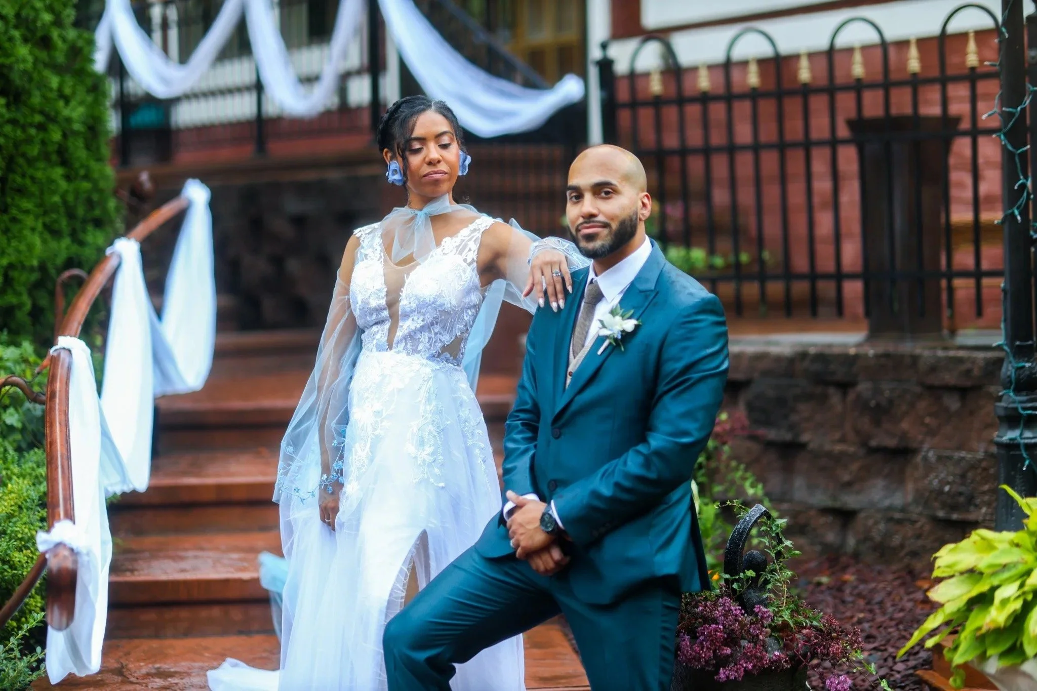 A bride and groom pose outdoors in front of a brick building, decorated with white and blue fabric, with greenery and flowers around them.