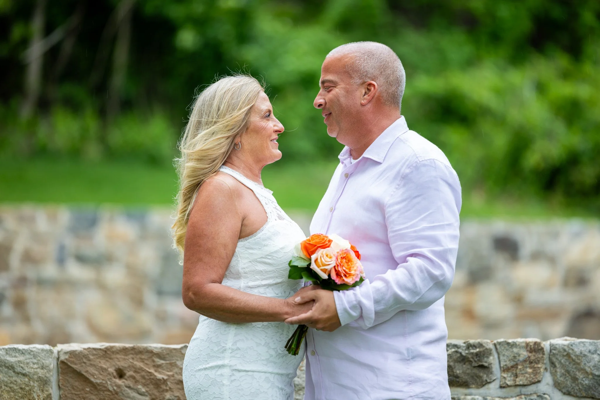 A couple standing outdoors, facing each other and smiling. The woman is holding a bouquet of flowers and is wearing a white lace dress. The man is wearing a white button-up shirt. Green trees and a stone wall are in the background.