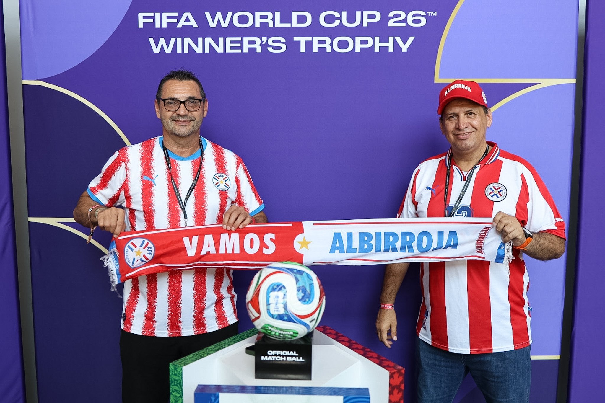 Two fans holding a team scarf beside a trophy display at a FIFA World Cup 26 brand activation, captured by David Pierre Studio