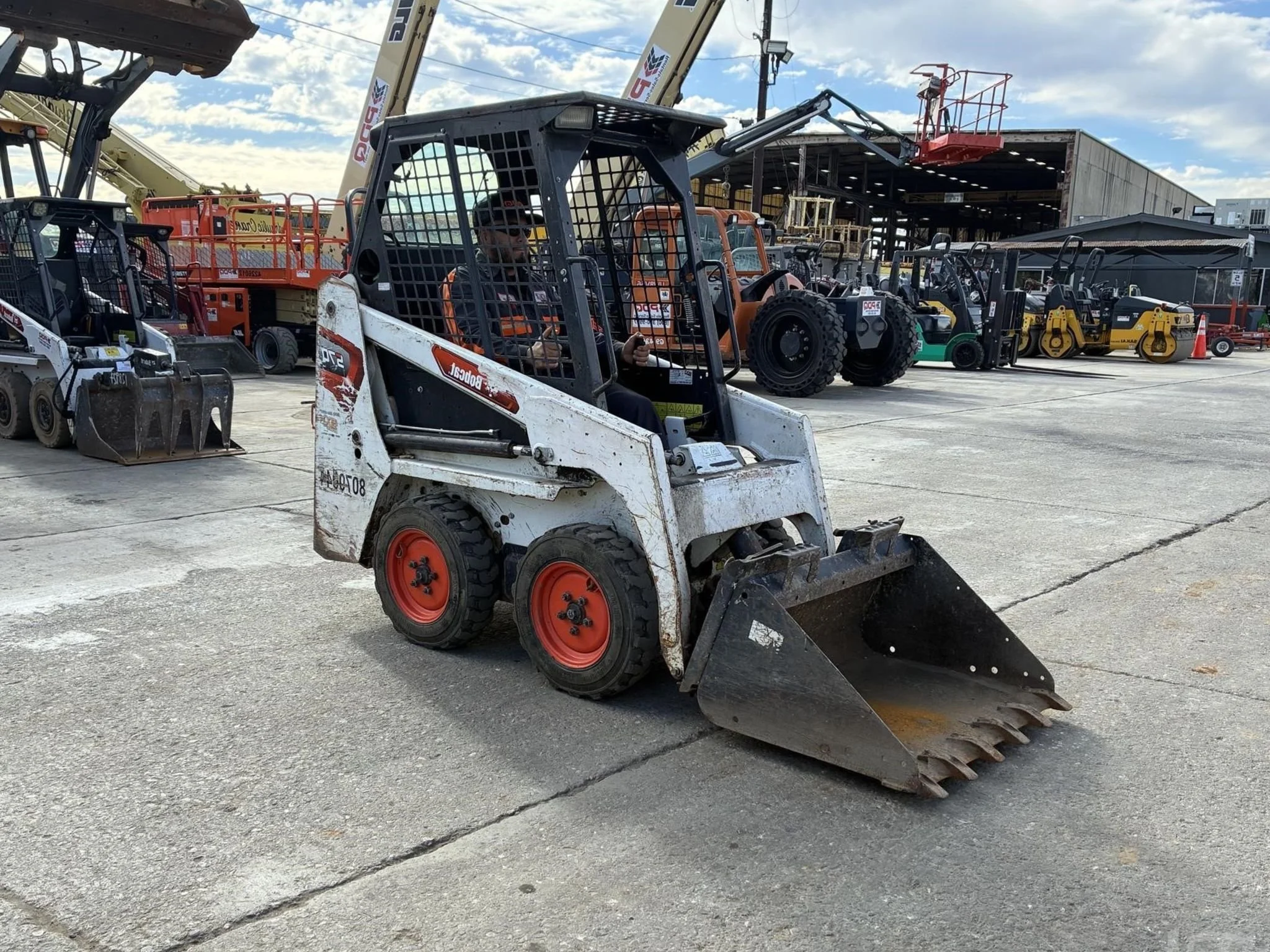 A skid-steer loader parked on a concrete surface with various construction equipment and machinery in the background. Available for rent in Santa Ana, CA