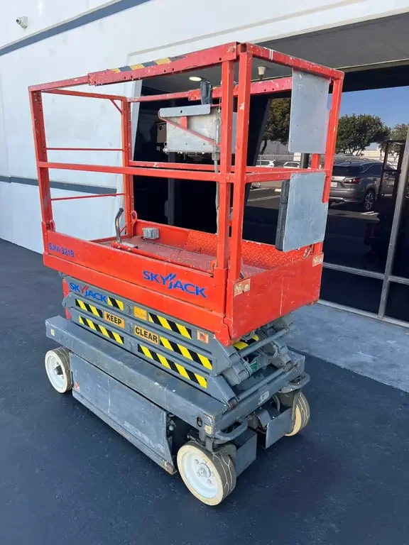 A red skyjack electric scissor lift parked outside a building with glass doors and windows.