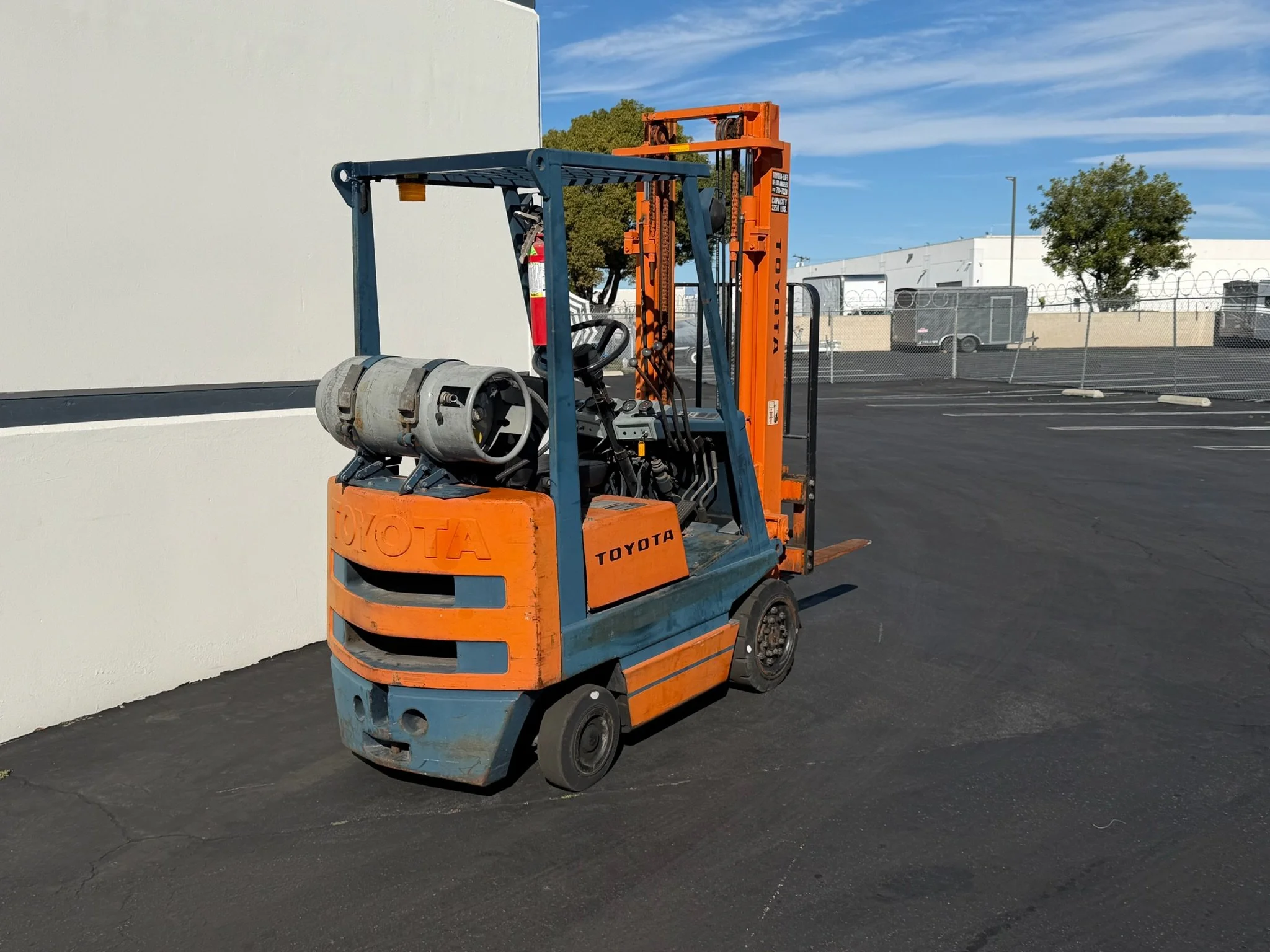 An orange and blue Toyota forklift parked on asphalt in a parking lot near a white wall, with a chain-link fence and buildings in the background. Forklift rentals in Santa Ana, CA