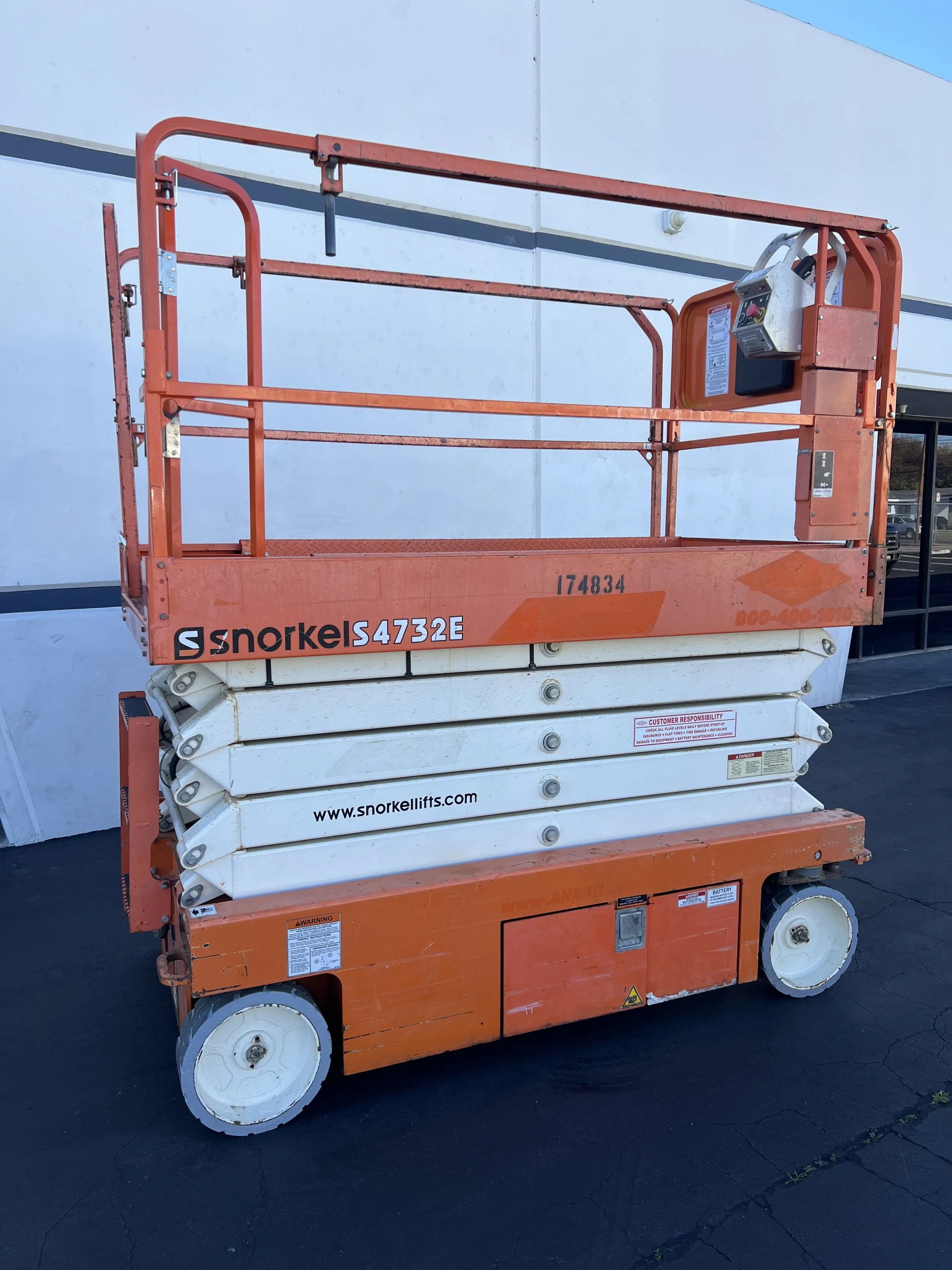 Orange and white scissor lift with the brand name Snorkel, parked on asphalt outside a building with a white wall and blue sky in the background. Available for rent in Santa Ana, CA