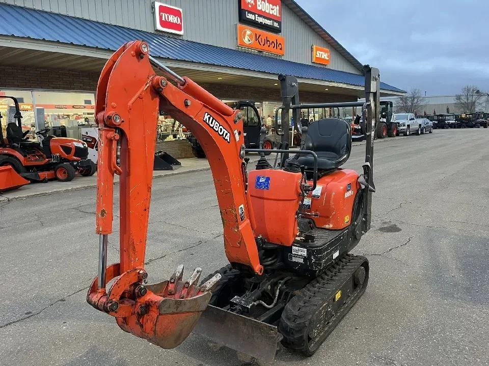 Orange Kubota mini excavator with black tracks parked outside a store with various outdoor power equipment and vehicles in the background. Available for rent in Santa Ana, CA