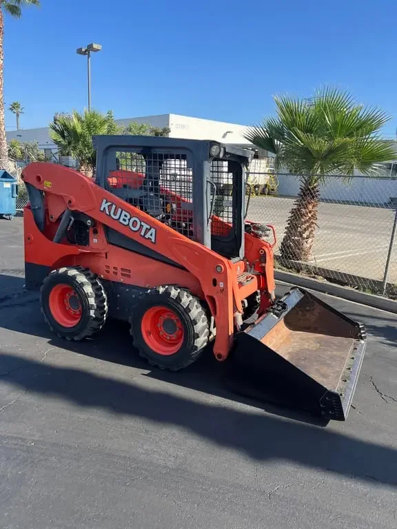 A small Kubota skid-steer loader parked on a paved area with a chain-link fence, palm trees, and a building in the background. Available for rent in Santa Ana, CA