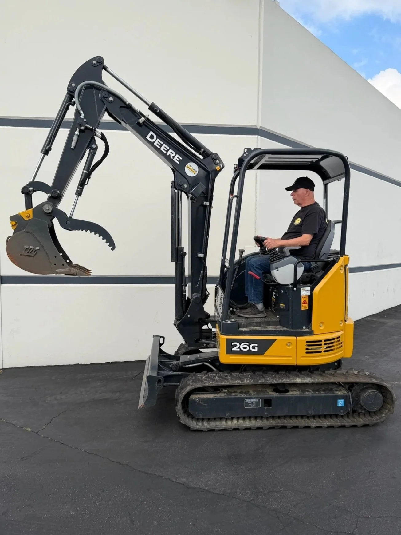 A man operating a yellow and black mini excavator with tracks, labeled 26G, in front of a white building with gray accents, on a black paved surface. Available for rent in Santa Ana, CA